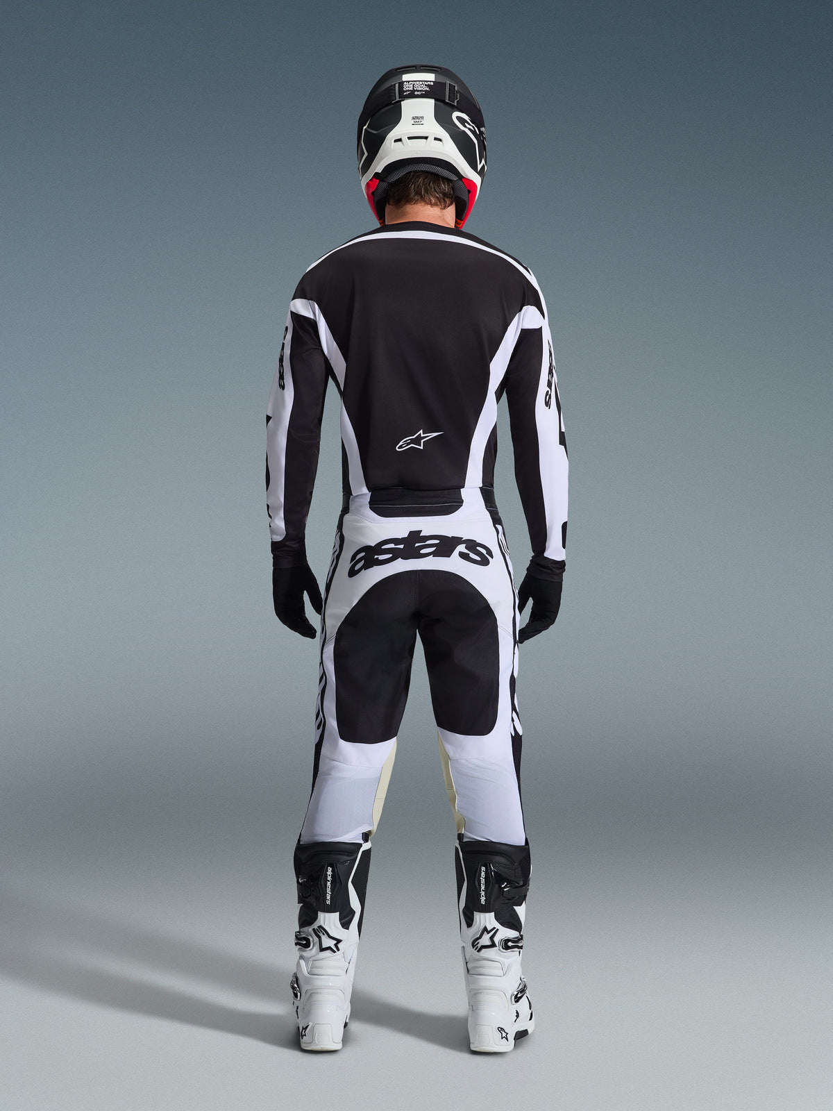 Vy bakifrån av en motocrossförare klädd i 2026 Racer Riway utrustningsset i svart och vitt, med en svart tröja med vita detaljer, vita byxor med framträdande 'astars'-logotyp på sätet, svart hjälm, vita stövlar och svarta handskar, komplett professionell racingutrustning för terrängkörning.