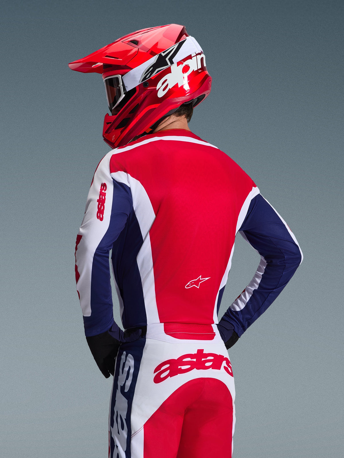 Motocrossförare sedd bakifrån iförd Alpinestars 2026 Racer Riway-tröja, färgställning i vitt, blått och rött, matchande offroad-byxor med tydlig astars-branding, röd hjälm med vit Alpinestars-logotyp, svarta handskar, koordinerad motocrossutrustning