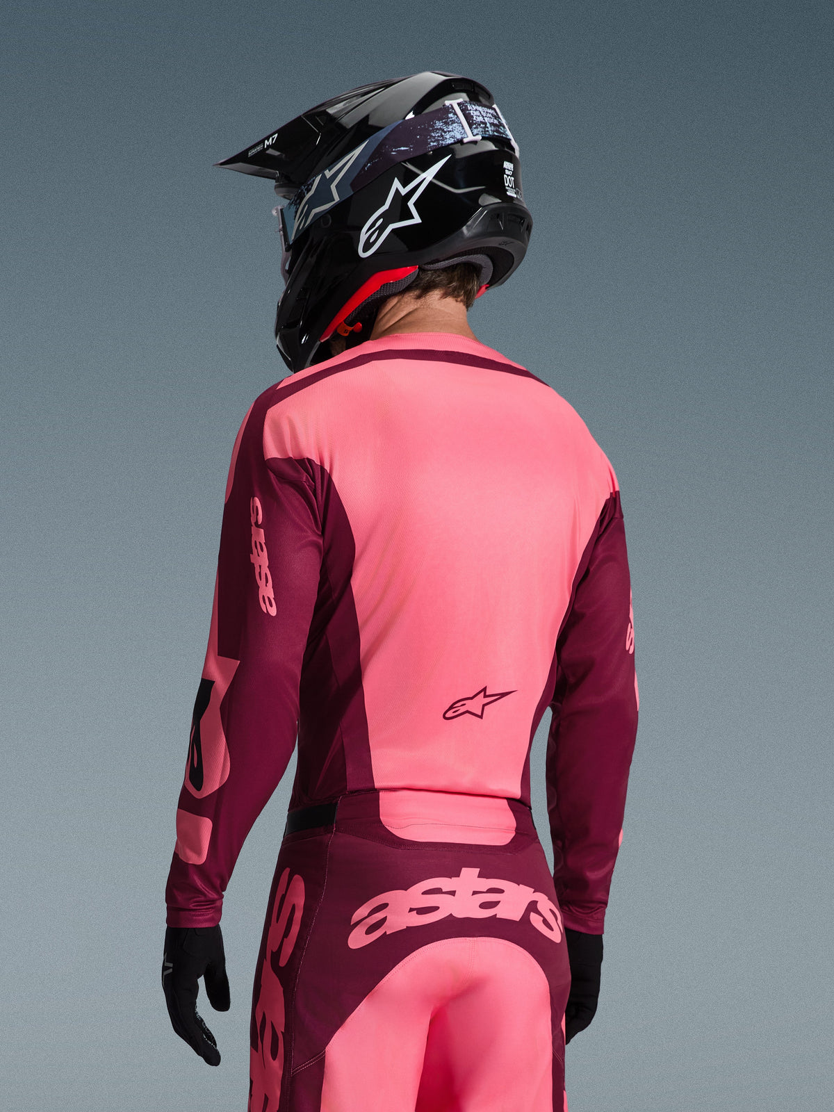 Motocrossförare sedd bakifrån i Alpinestars 2026 Racer Riway Gear Set, ljusrosa och vinröd tröja och byxor, svart motocrosshjälm med vit logotyp och svarta handskar, med 'astars'-branding på ryggen.