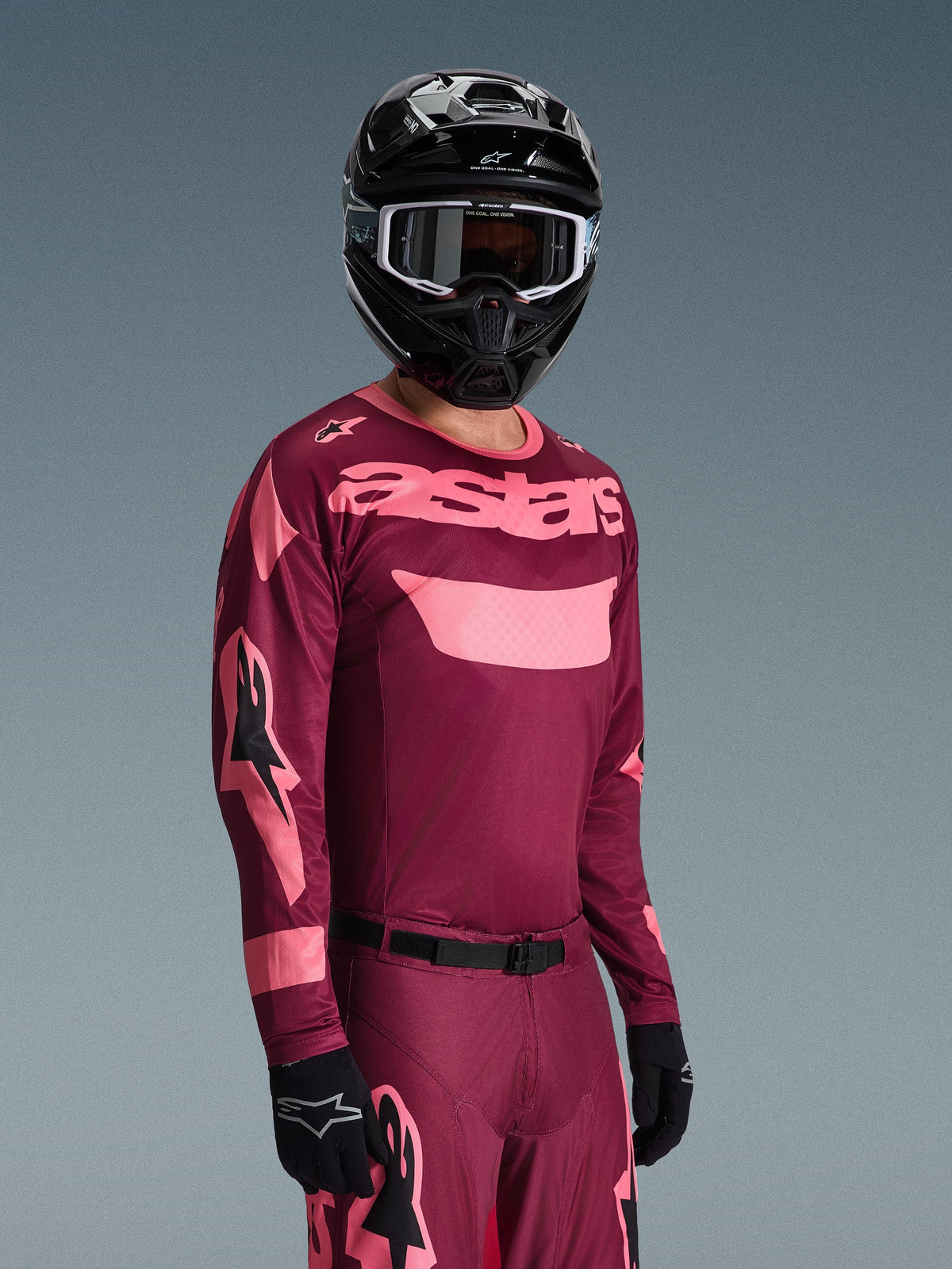 Motocrossförare i Alpinestars 2026 Racer Riway-tröja i färgen Dark Red Coral med matchande byxor, med korallrosa 'astars'-logotyp på bröstet, kombinerat med en svart hjälm och svarta handskar, koordinerat set med racingutrustning för terrängkörning.