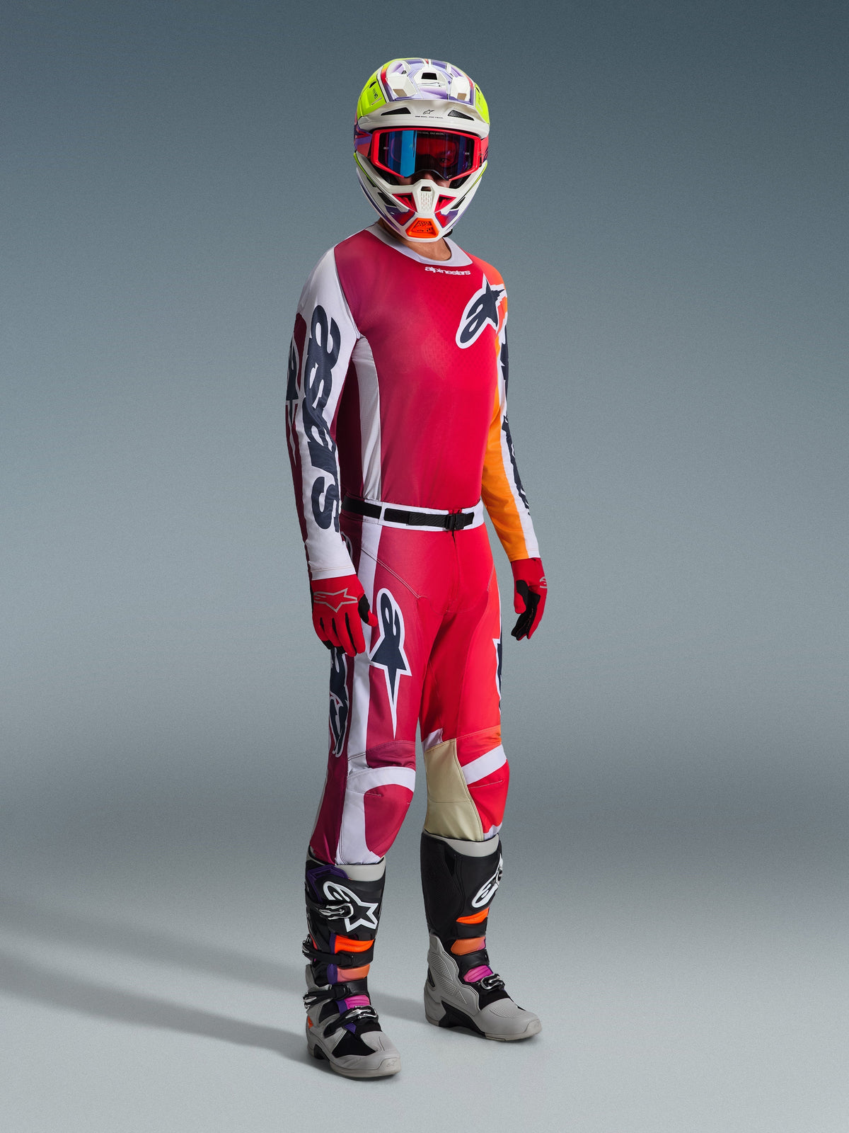 Motocrossförare i komplett tävlingsutrustning, Alpinestars 2026 Racer Portl-tröja i rött och ljusgrått, matchande motocrossbyxor med vita och mörkgröna detaljer, flerfärgad hjälm med röda goggles, grå och svarta stövlar med orange och lila detaljer, röda handskar, koordinerad outfit för terrängkörning