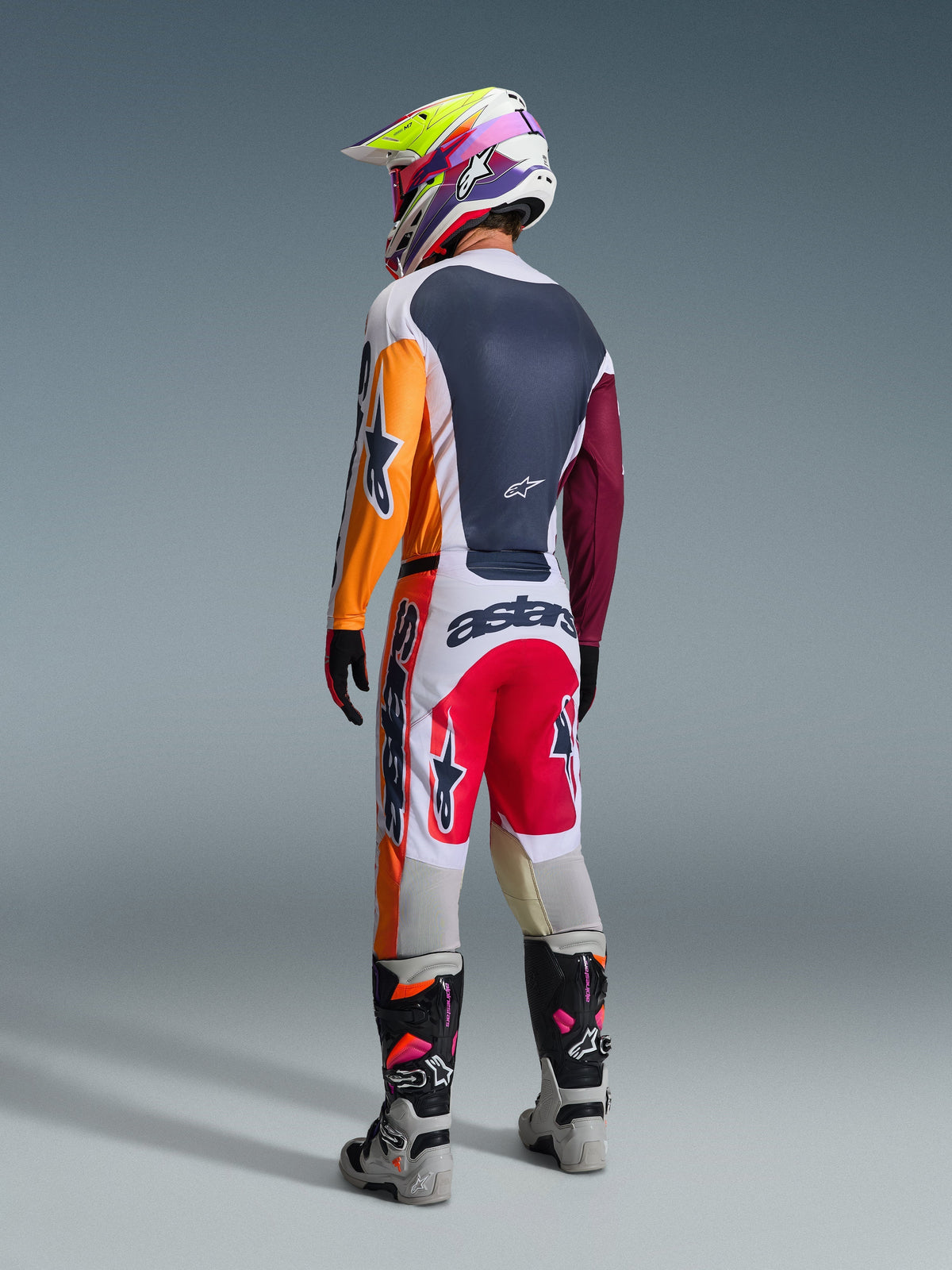 Motocrossförare i komplett racingutrustning sedd bakifrån, Alpinestars 2026 Racer Portl-tröja i Red Light Gray och Dark Green med orangea och vinröda paneler, matchande vita och röda byxor, grå och svarta stövlar med rosa detaljer, flerfärgad hjälm, koordinerad outfit för terrängkörning.