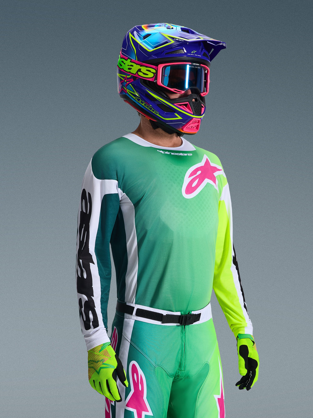 Alpinestars 2026 Racer Portl tröja, motocrosströja, grön, vit och svart, bärs av en förare med matchande gröna byxor, fluorescerande handskar och en färgstark blå och lila hjälm med rosa goggles, med en rosa Astars-logotyp på bröstet.