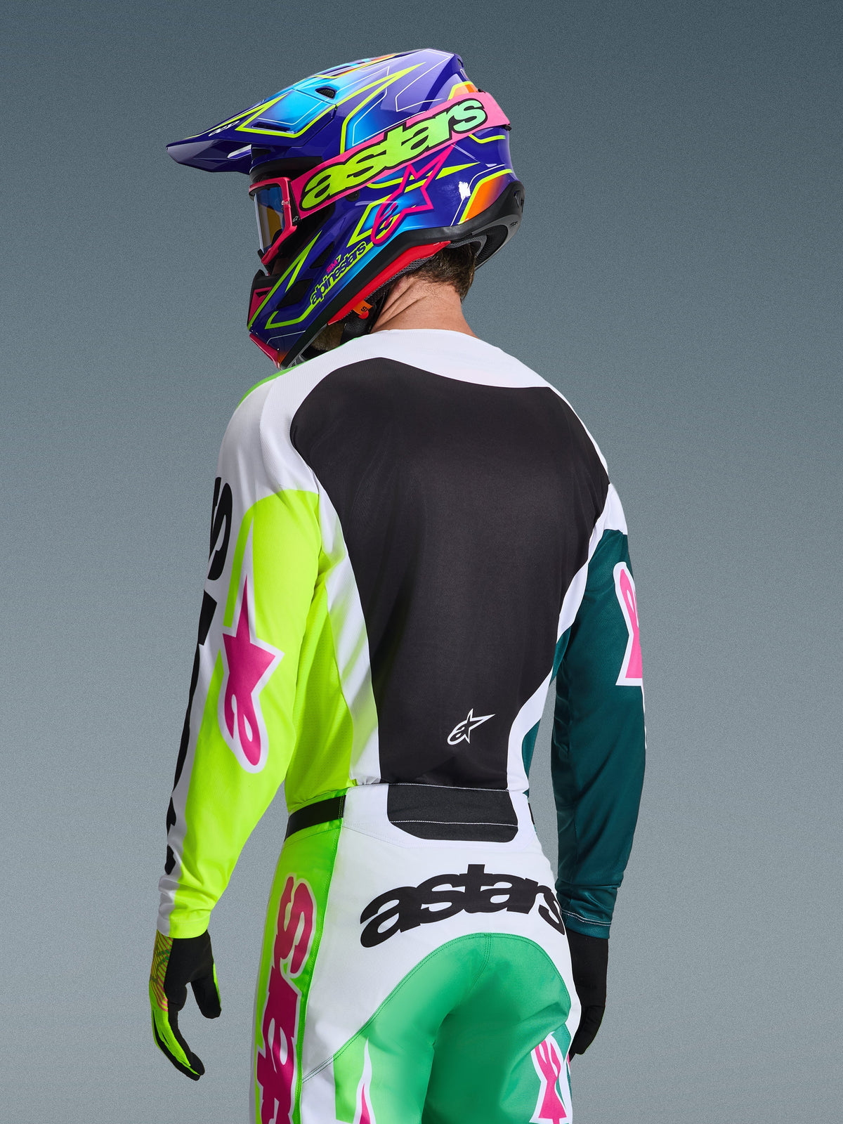 Motocrossförare sedd bakifrån i Alpinestars 2026 Racer Portl-tröja i Green White Black, med rosa stjärnlogotyper, matchande byxor och en färgglad blå och lila hjälm, koordinerad racingutrustning för offroad-prestanda.