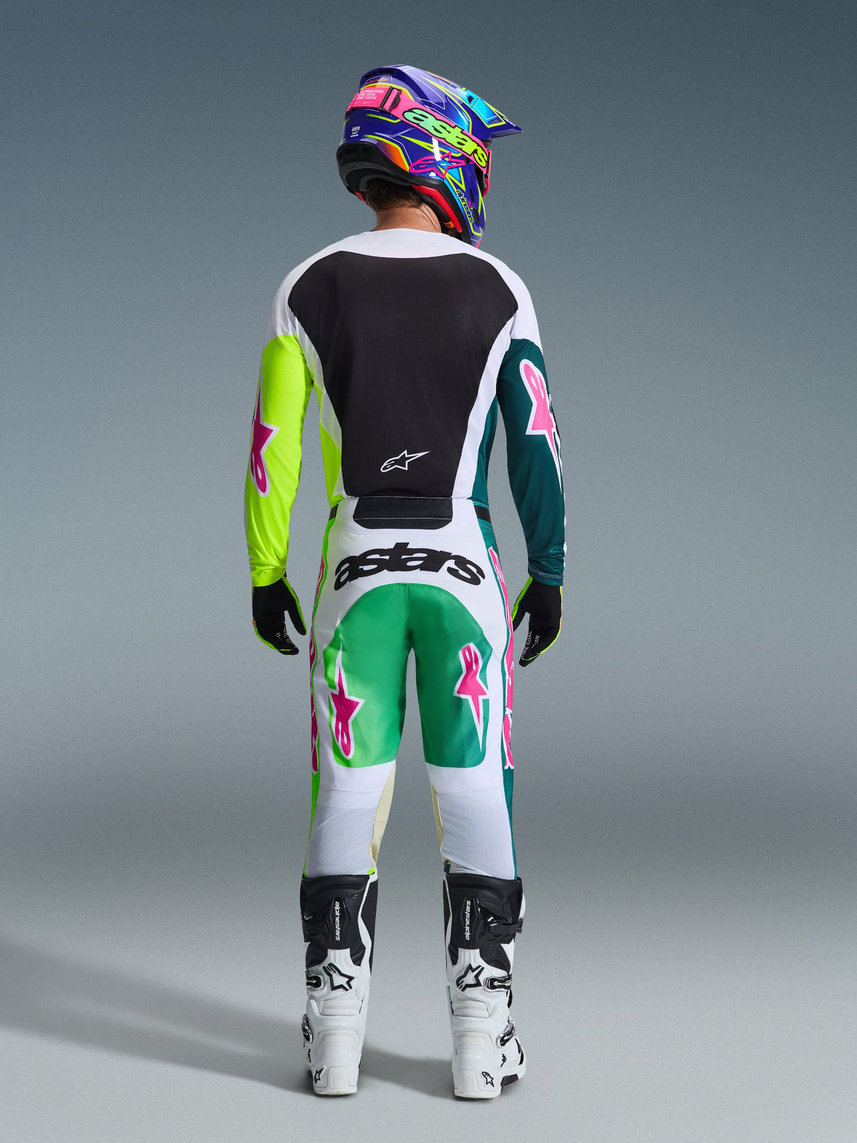 Rear view of motocross rider wearing the Alpinestars 2026 Racer Portl Gear Set, white jersey with a black back panel and neon yellow and dark green sleeves featuring pink stars, matching white pants with green and teal accents and pink star logos, coordinated with a colorful helmet, black gloves, and white and black boots.
