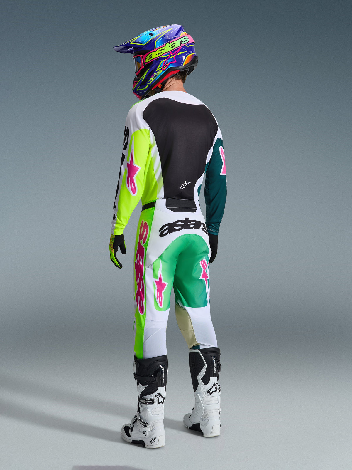 Motocross rider from behind wearing the Alpinestars 2026 Racer Portl Gear Set, jersey featuring neon yellow and teal sleeves with pink logos, matching white and green pants with bold pink accents, colorful helmet and white boots, professional off-road racing gear