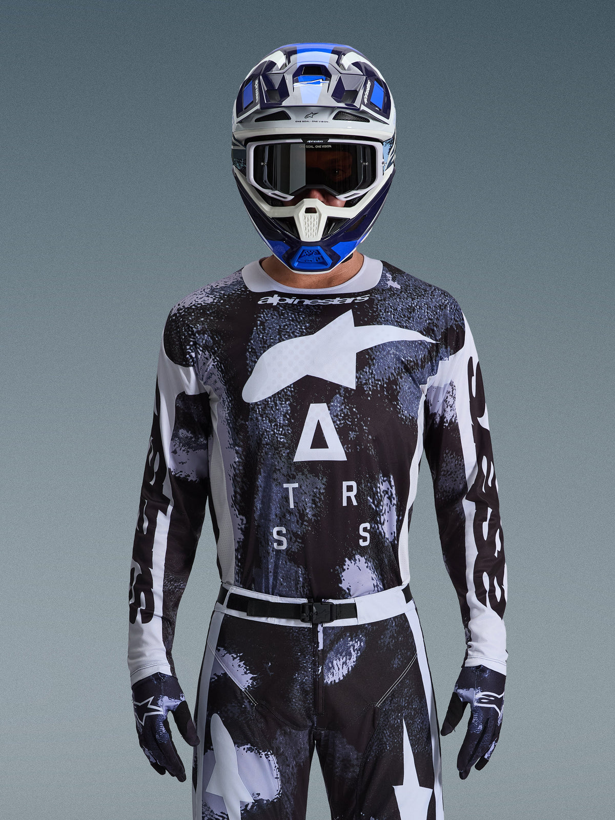Motocross rider wearing the Alpinestars 2026 Racer Lahnd Gear Set, black and white abstract camo jersey with large white logos and "ATRS" lettering, matching pants and gloves, blue and white helmet with goggles, high-performance racing outfit for off-road motocross