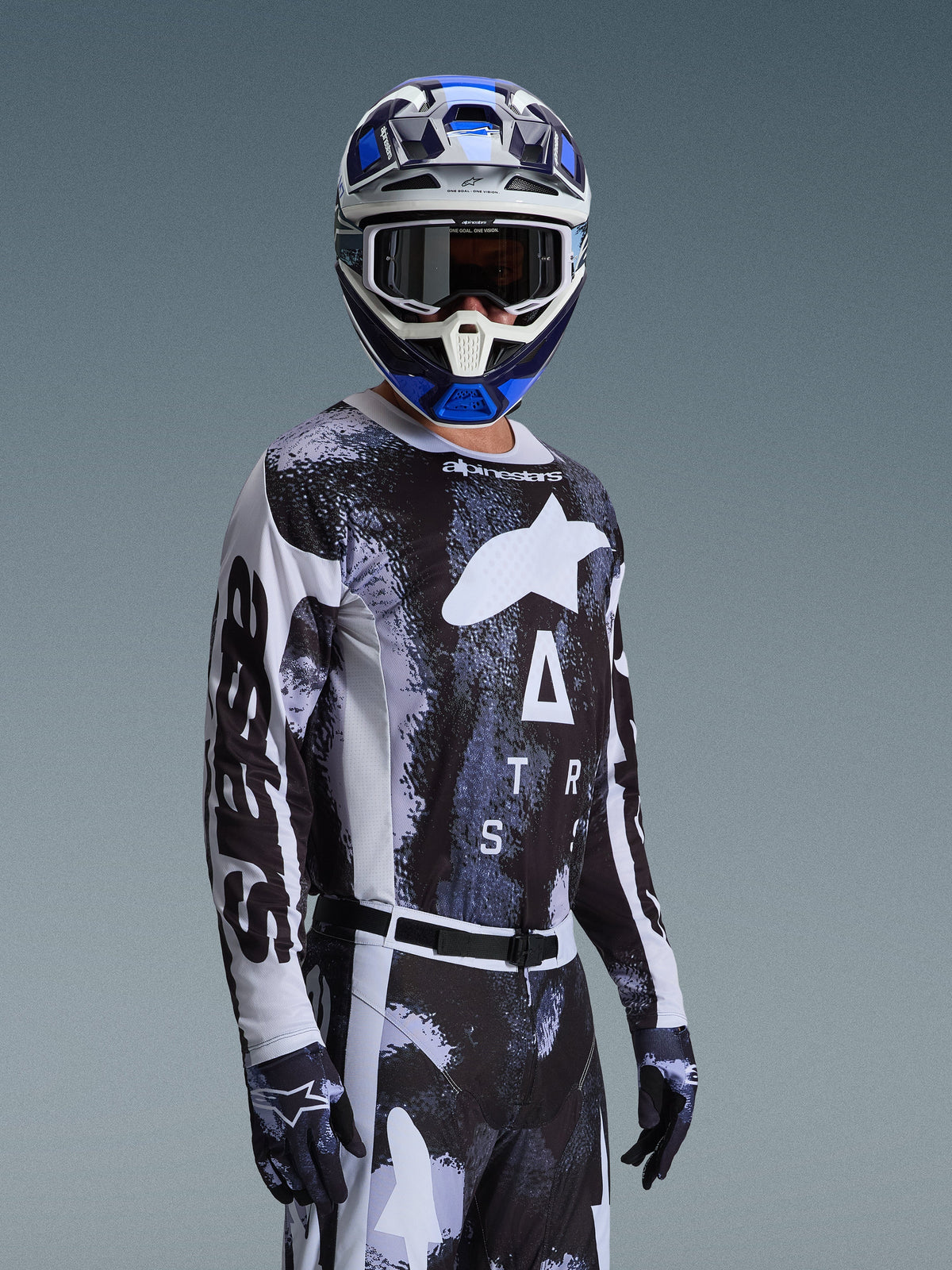 Motocross rider wearing the Alpinestars 2026 Racer Lahnd Jersey in Iron Camo, featuring a black and grey camouflage pattern with a large white Alpinestars logo on the chest, styled with a matching helmet and gloves for off-road racing.