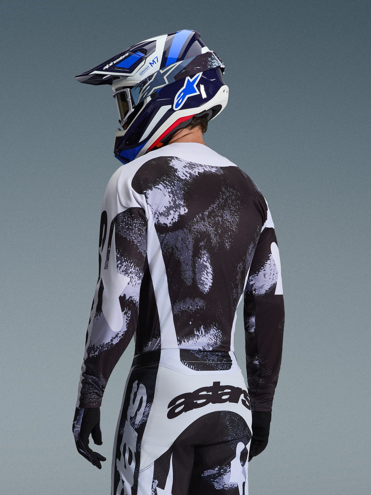 Alpinestars 2026 Racer Lahnd Gear Set, Motocross rider from behind wearing jersey and pants with black and white speckled graphic, paired with a blue and white Alpinestars helmet and black gloves, performance off-road racing gear