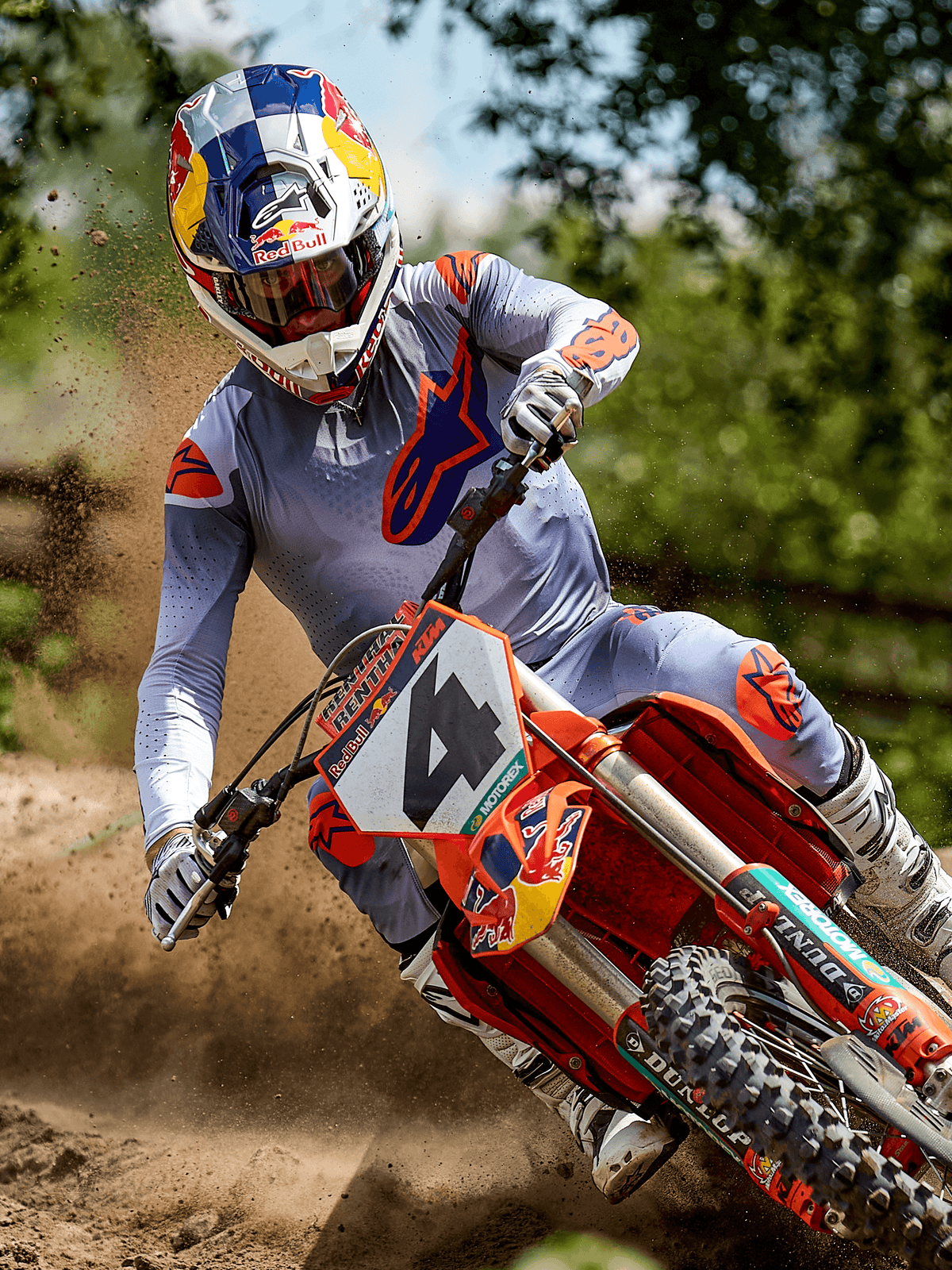 Motocrossförare i aktion på en jordbana, bär Alpinestars 2025 Supertech Lipan Jersey i Gray Orange Fluo, matchande grå byxor med orange detaljer, vita stövlar och en Red Bull-märkt hjälm, tar en kurva på en orange motocrosscykel med jordstänk, professionell racingutrustning för offroad