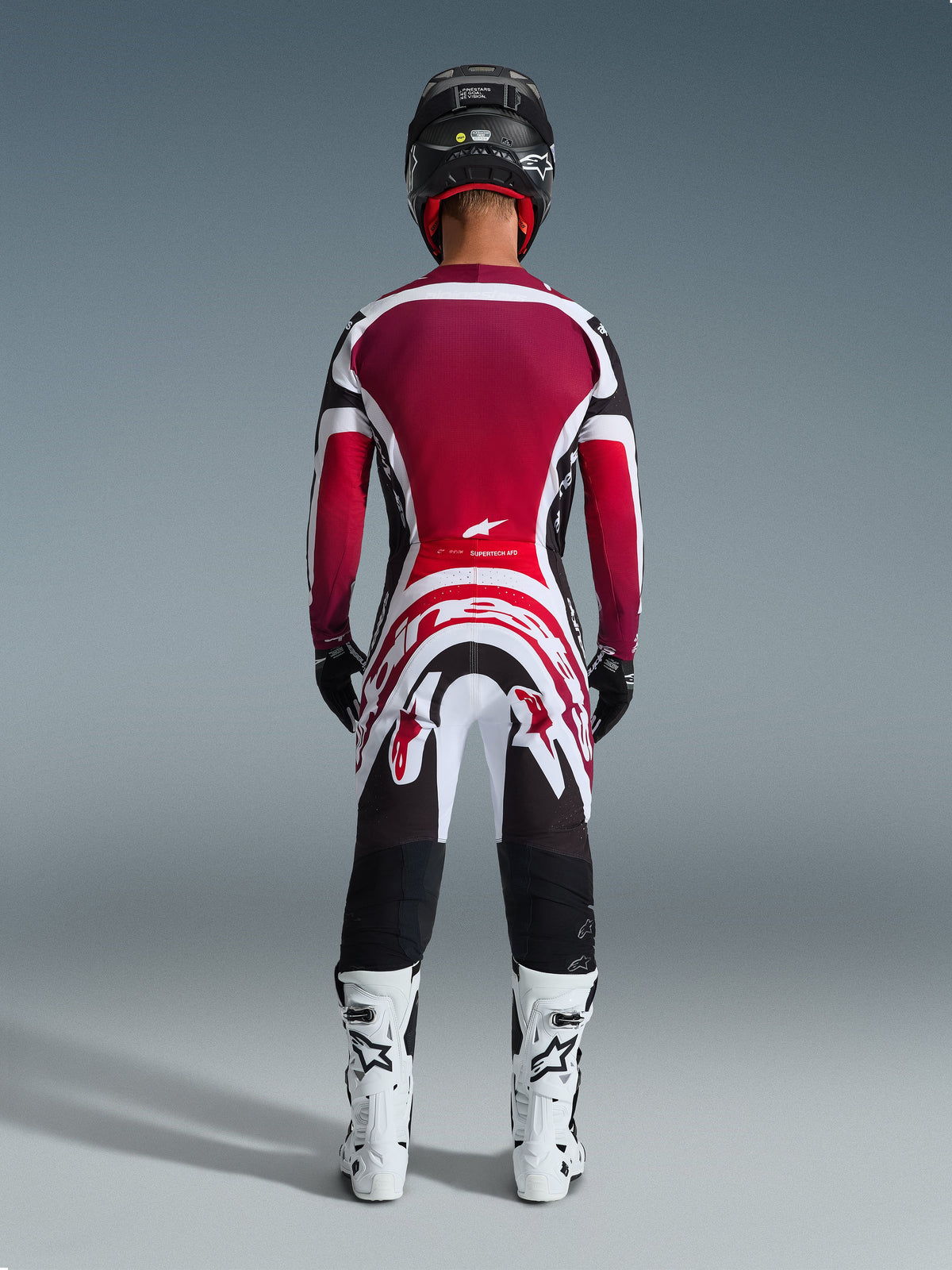 Alpinestars 2026 Supertech AFD Vista utrustningsset, vy bakifrån av en motocrossförare i professionell racingutrustning, vinröd och röd tröja med vita och svarta detaljer, matchande byxor med kraftfull grafik, vita stövlar och svart hjälm, koordinerad motocrossoutfit.