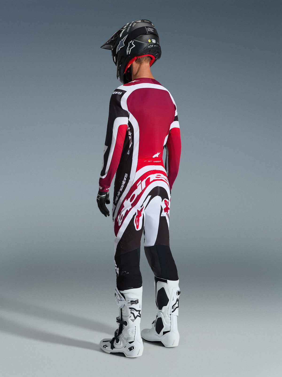 Motocrossförare sedd bakifrån i Alpinestars 2026 Supertech AFD Vista-utrustningsset, röd, vit och svart tröja och byxor med framträdande Alpinestars-logotyper, svart hjälm, vita och svarta stövlar och svarta handskar för en komplett professionell racinglook.