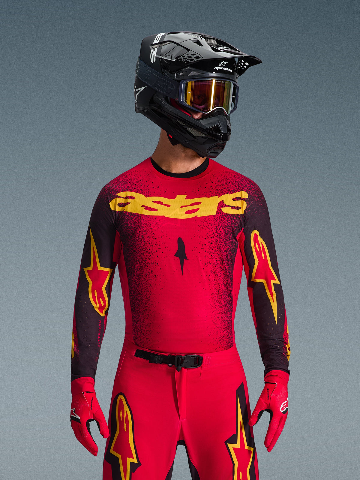 Alpinestars 2026 Supertech Scenz Gear Set, Motocross rider wearing a red jersey with yellow 'astars' branding and black speckled details, red pants with yellow star logos on the thighs, red racing gloves, and a black helmet with reflective yellow goggles, pro-level racing outfit for off-road performance