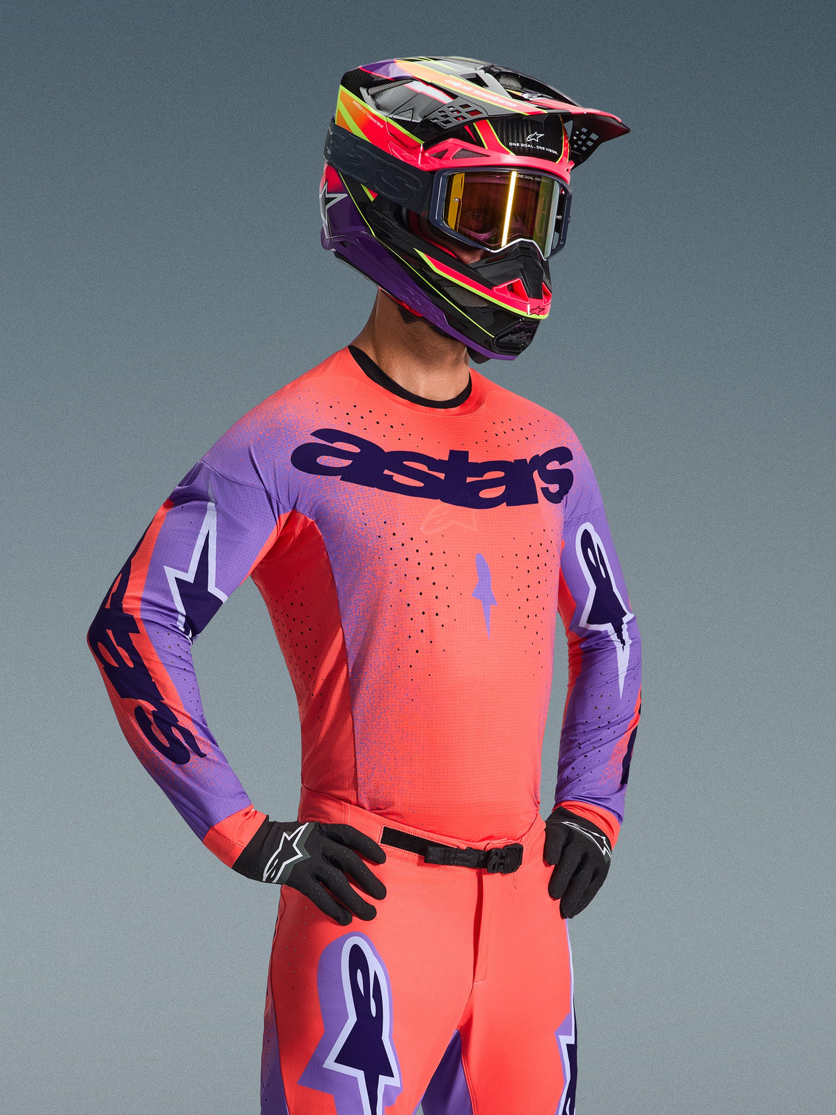 Motocrossförare i Alpinestars 2026 Supertech Scenz Jersey, Hot Coral och lila med framträdande 'astars'-logotyp på bröstet, matchande korallfärgade byxor och koordinerad lila och korallfärgad hjälm, laserskurna perforeringar för högpresterande terrängkörning