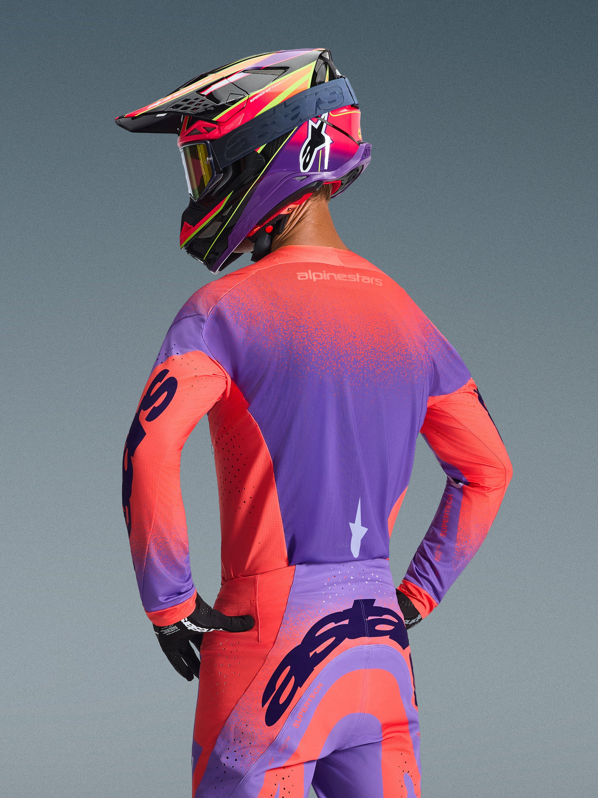 Motocrossförare sedd bakifrån i 2026 Alpinestars Supertech Scenz-tröja i Hot Coral och lila, med en levande spräcklig gradientdesign, matchande motocrossbyxor och en färgglad racinghjälm, professionell utrustning för terrängprestanda.