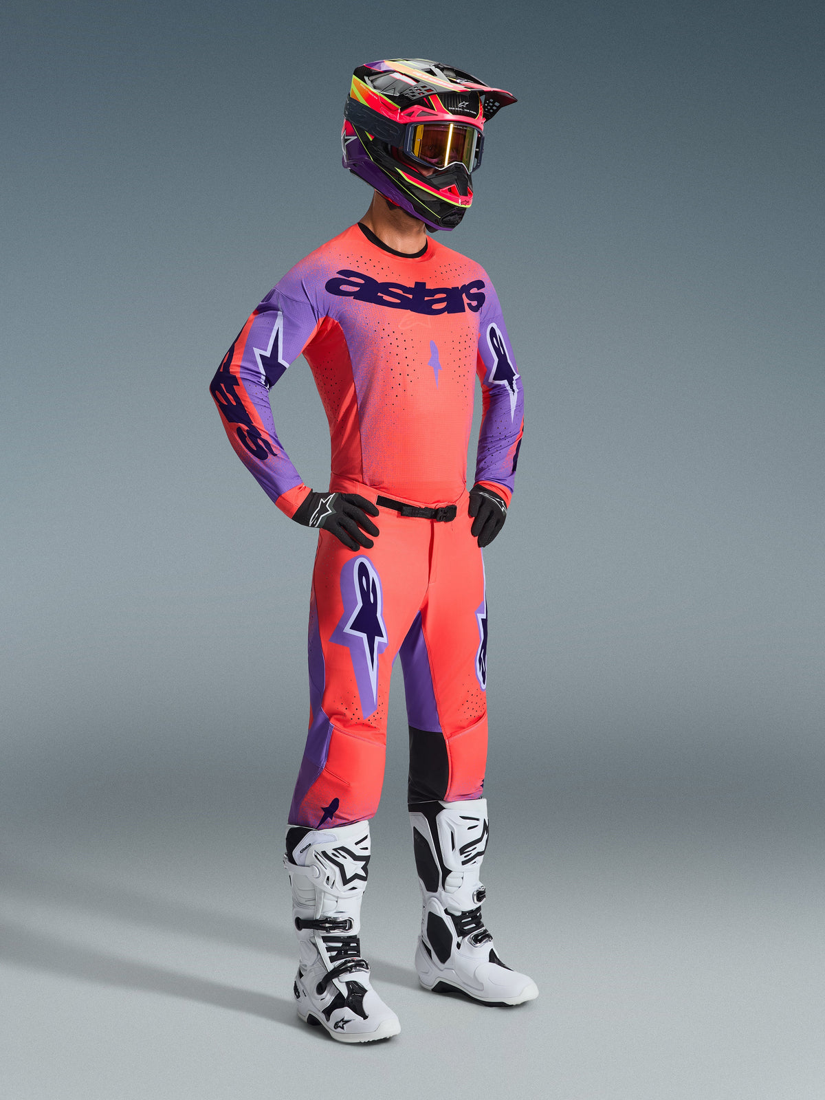 Motocrossförare klädd i komplett Alpinestars 2026 Supertech Scenz utrustningsset, Hot Coral-tröja med lila ärmar och 'astars'-logotyp, matchande korallfärgade byxor med lila detaljer, flerfärgad hjälm med guldtonade goggles, vita och svarta stövlar, svarta handskar, koordinerad racingoutfit på proffsnivå för terrängkörning