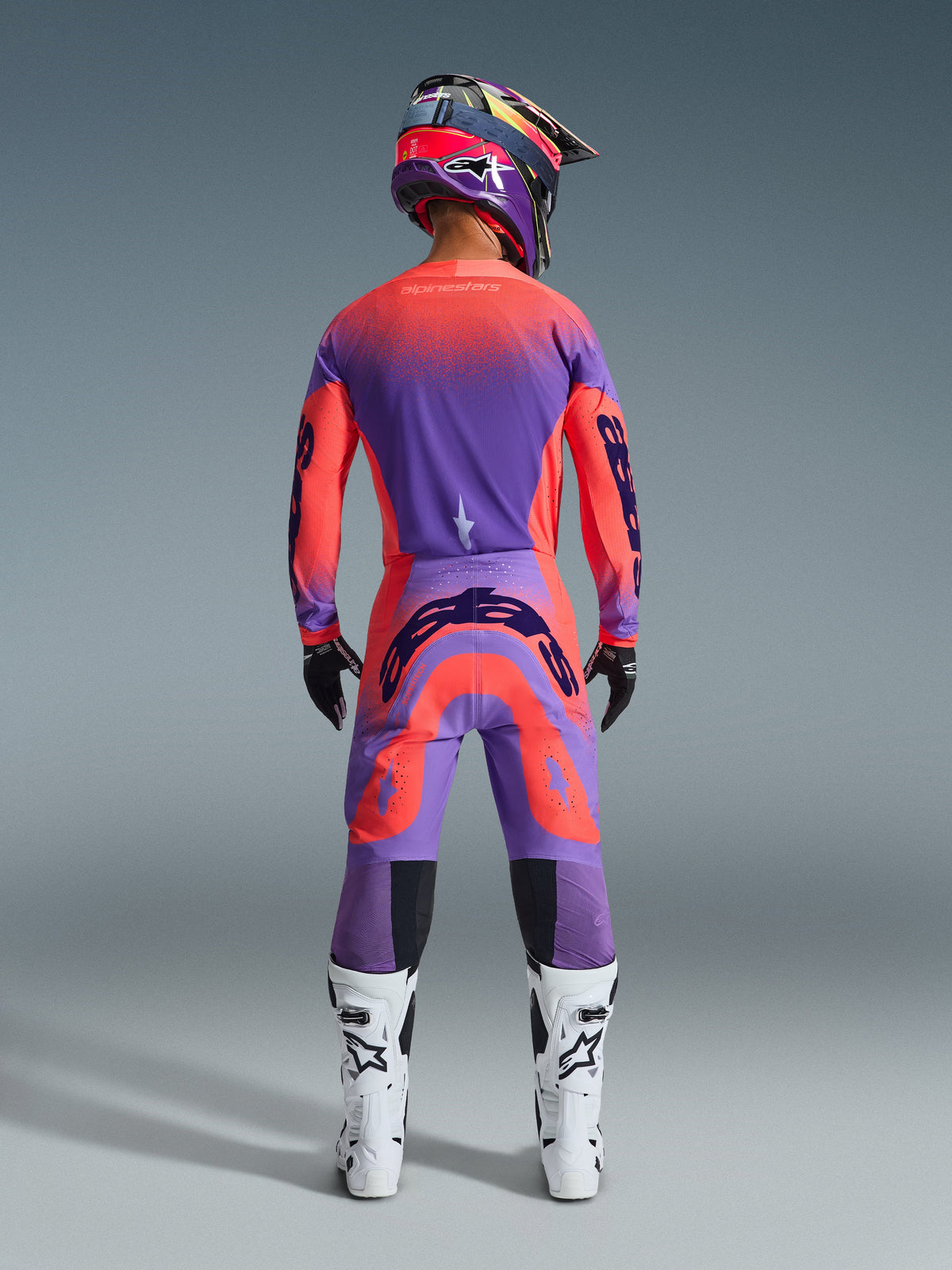 Motocrossförare sedd bakifrån bärandes 2026 Supertech Scenz-utrustningsset i Hot Coral Purple, färgstark tröja och byxor med tonade övergångar, Alpinestars-logotyper, matchande hjälm, vita och svarta stövlar samt svarta handskar, professionell utrustning för terrängkörning på elitnivå.