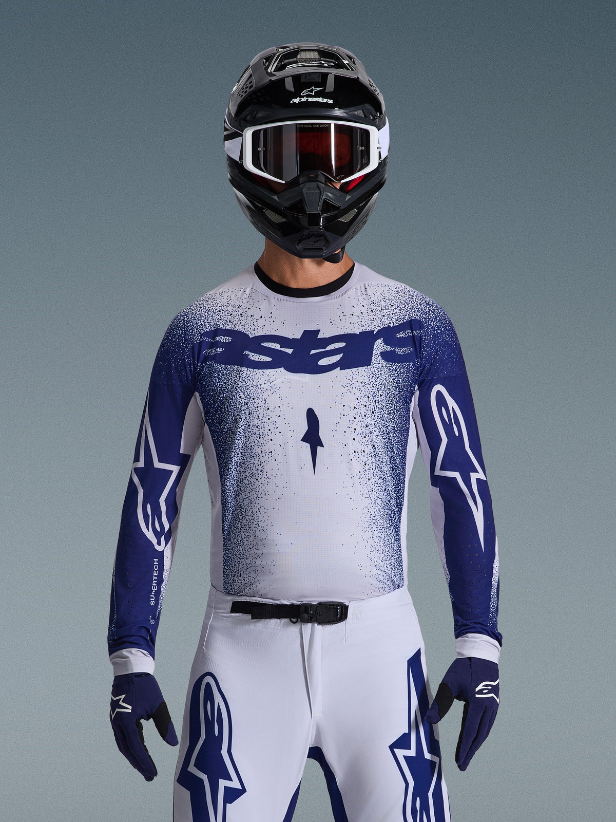 Motocrossförare i Alpinestars 2026 Supertech Scenz-utrustningsset, vit tröja med blått spräckligt tonat mönster och 'astars'-logotyp, matchande vita byxor med blå detaljer, svart hjälm med vita goggles och blå handskar, atletisk racingpassform på proffsnivå.