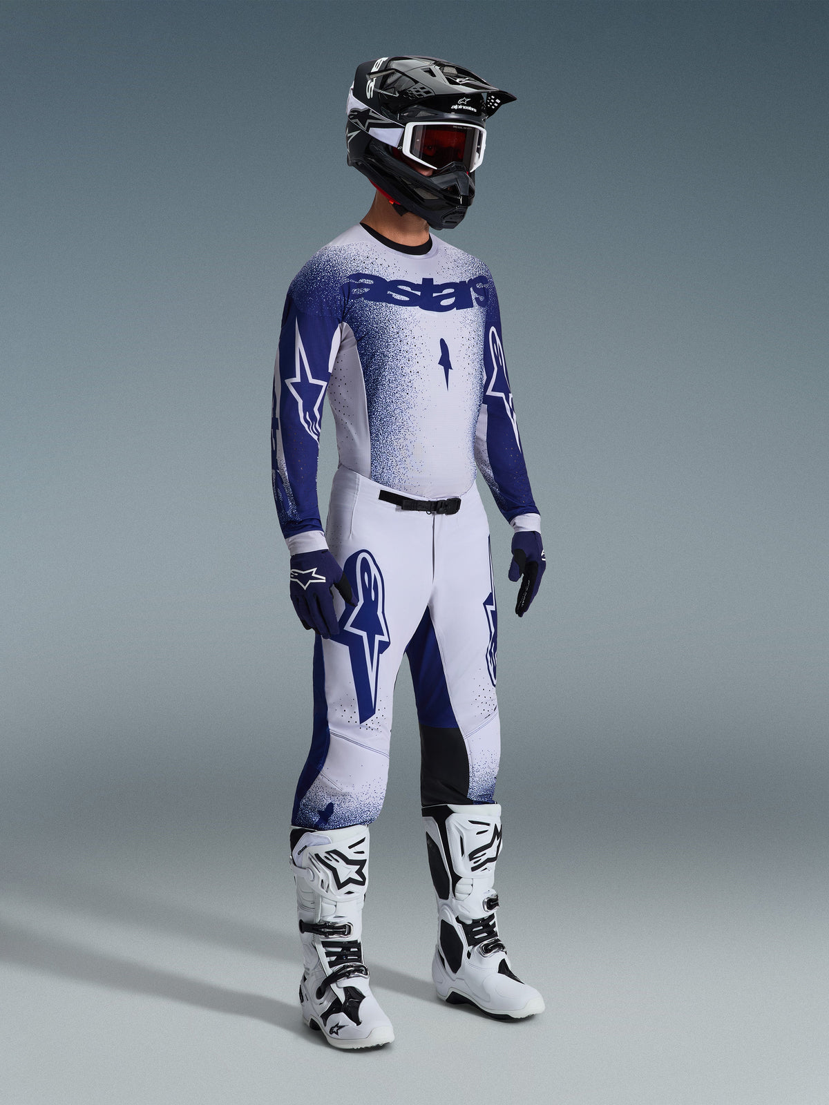 Alpinestars 2026 Supertech Scenz-utrustningsset, motocrossförare i vit och marinblå racingtröja och byxor med en tonad stipple-effekt, framträdande Astars-logotyp, kombinerat med svart och vit hjälm, marinblå handskar och vita motocross-stövlar.