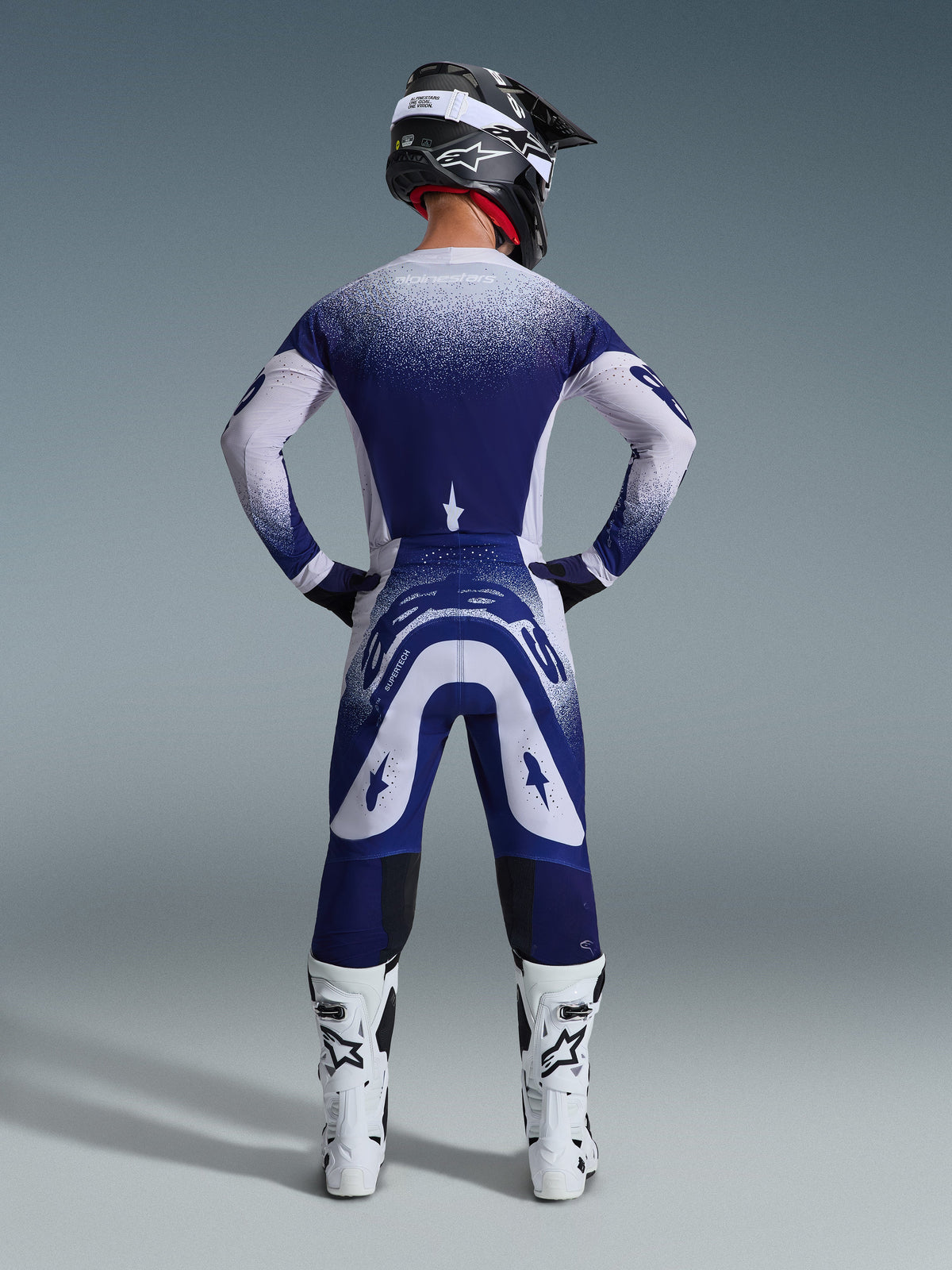 Alpinestars 2026 Supertech Scenz utrustningsset, vy bakifrån av en motocrossförare i blå och vit racingtröja och byxor, prickig gradienteffekt, bärs med vita motocross-stövlar och svart hjälm, motocrossutrustning på proffsnivå