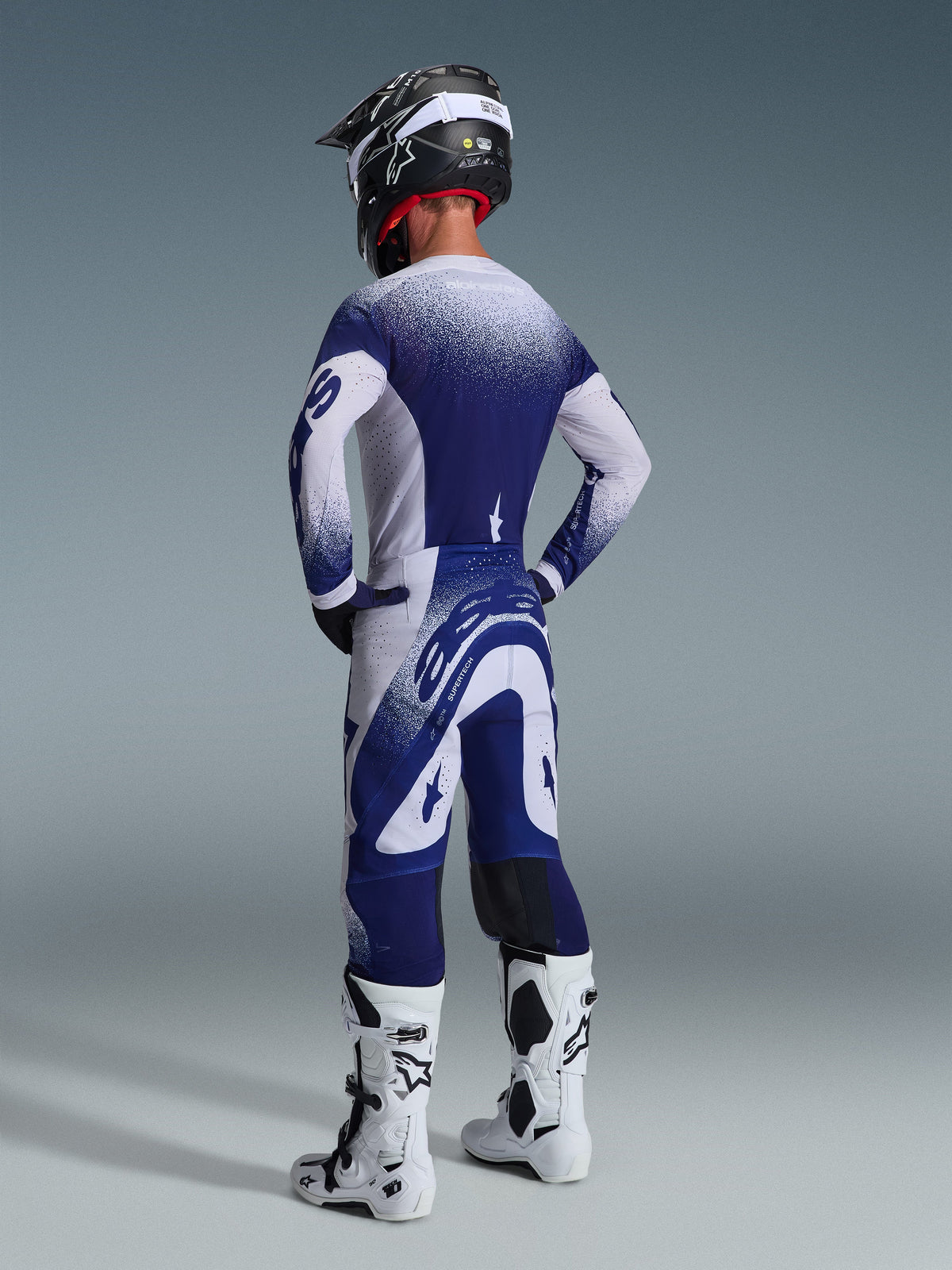 Motocrossförare klädd i Alpinestars 2026 Supertech Scenz-utrustningsset, marinblå och vit tröja och byxor med en spräcklig gradienteffekt, vy bakifrån som visar atletisk passform, kombinerat med vita och svarta stövlar och en svart hjälm, professionell racingutrustning för terrängprestanda