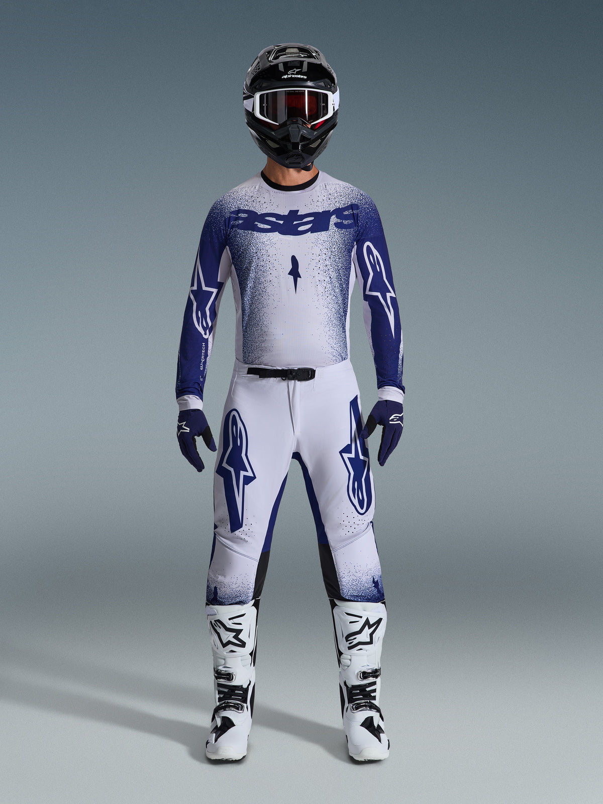 Motocrossförare i komplett racingutrustning, 2026 Supertech Scenz-tröja i ljusgrå och mörkblå, matchande ljusgrå byxor med blå logotyper, svart och vit hjälm med röda goggles, vita och svarta stövlar, mörkblå handskar, koordinerad outfit för terrängkörning