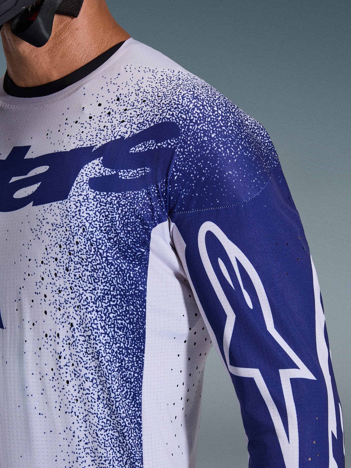 Alpinestars 2026 Supertech Scenz Tröja, ljusgrå och mörkblå, närbild på axel och bröst som visar laserskurna perforeringar, vit Astars-logotyp på blå ärm, spräcklig grafisk design, professionell motocrossutrustning.