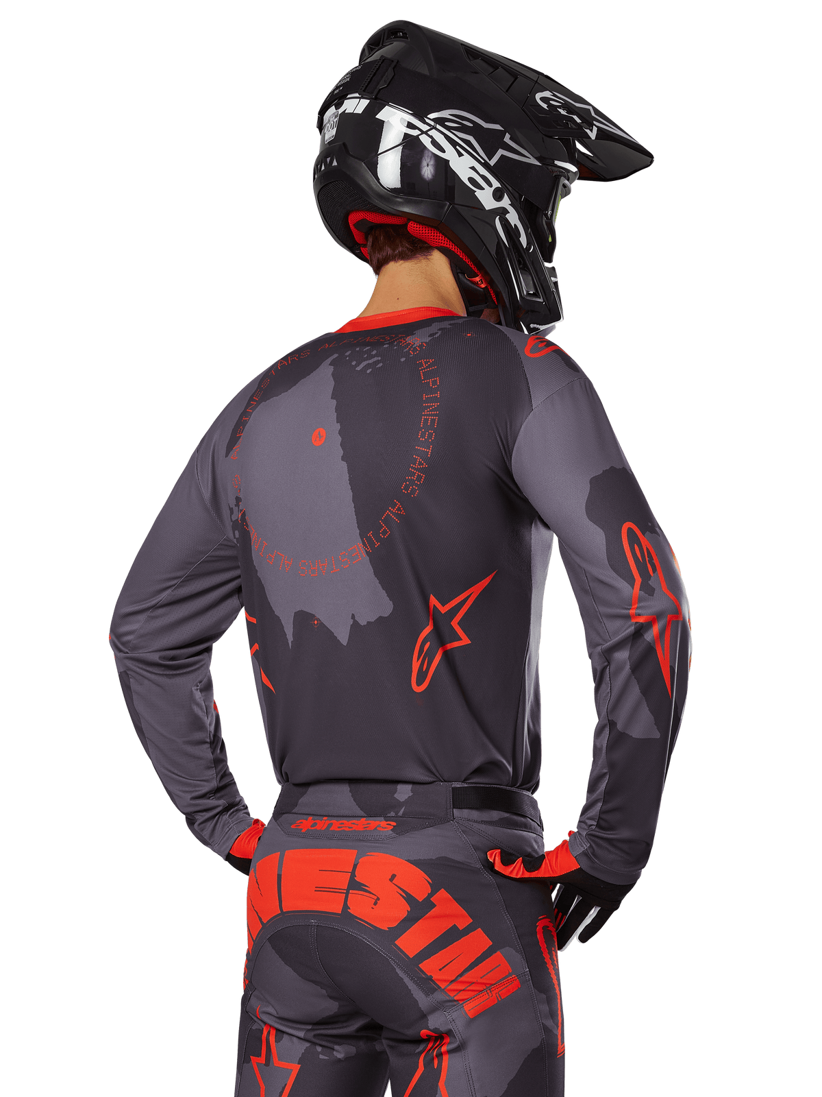 Alpinestars 2025 Racer Hollow-tröja, ryggvy av en motocrossförare i Gray Camo och Orange Fluo-utrustning, matchande kamouflagebyxor med stora orangea Alpinestars-bokstäver, svart hjälm, professionell design för terrängkläder