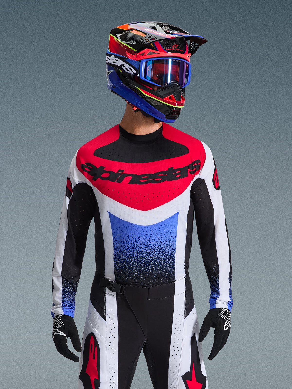 Alpinestars 2026 Techstar Knif tröja, motocrosströja, röd, vit och blå, med en framträdande bröstlogotyp och tonad färgövergång på bålen, bärs av en förare med en matchande flerfärgad hjälm, blå goggles och svarta handskar, professionell utrustning för terrängracing.