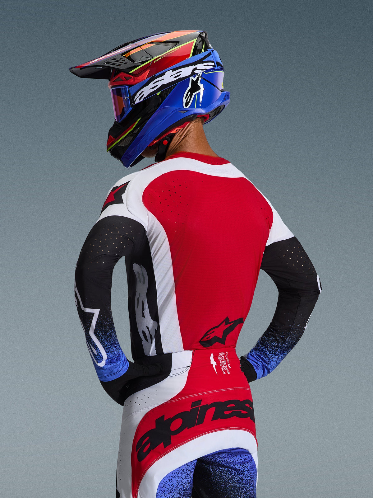 Motocrossförare sedd bakifrån bärandes 2026 Techstar Knif Jersey i rött, vitt och svart, med laserutskurna perforeringar på ryggen och framträdande logotyper, matchad med tillhörande byxor med blå detaljer och en blå och svart Alpinestars-hjälm, professionell offroad-racingestetik