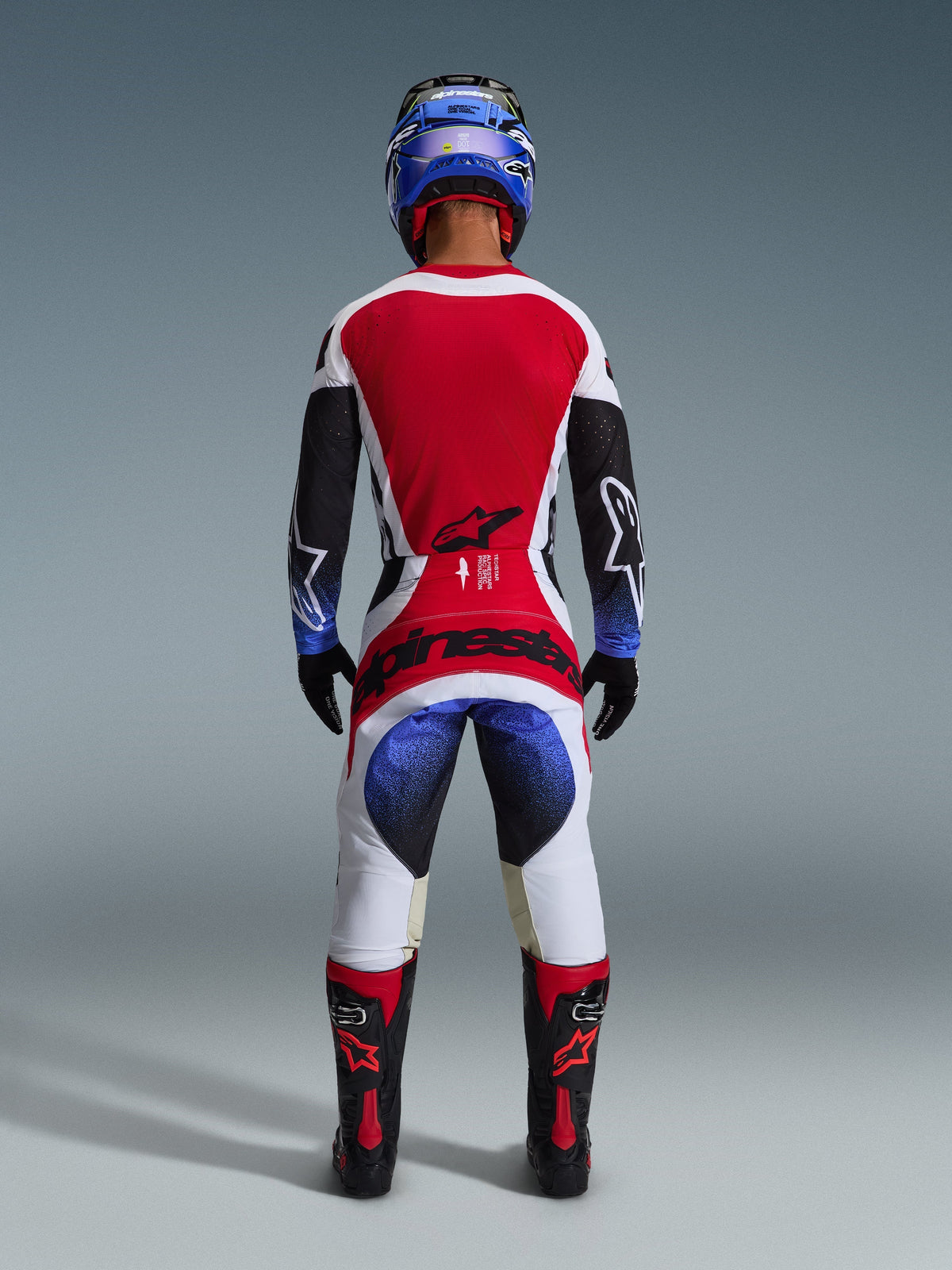 Motocross rider wearing the Alpinestars 2026 Techstar Knif Gear Set, viewed from behind, featuring a red white and black jersey with blue gradient accents on the sleeves, matching white and black pants with bold red branding, blue helmet, and black and red boots, professional off-road racing outfit.