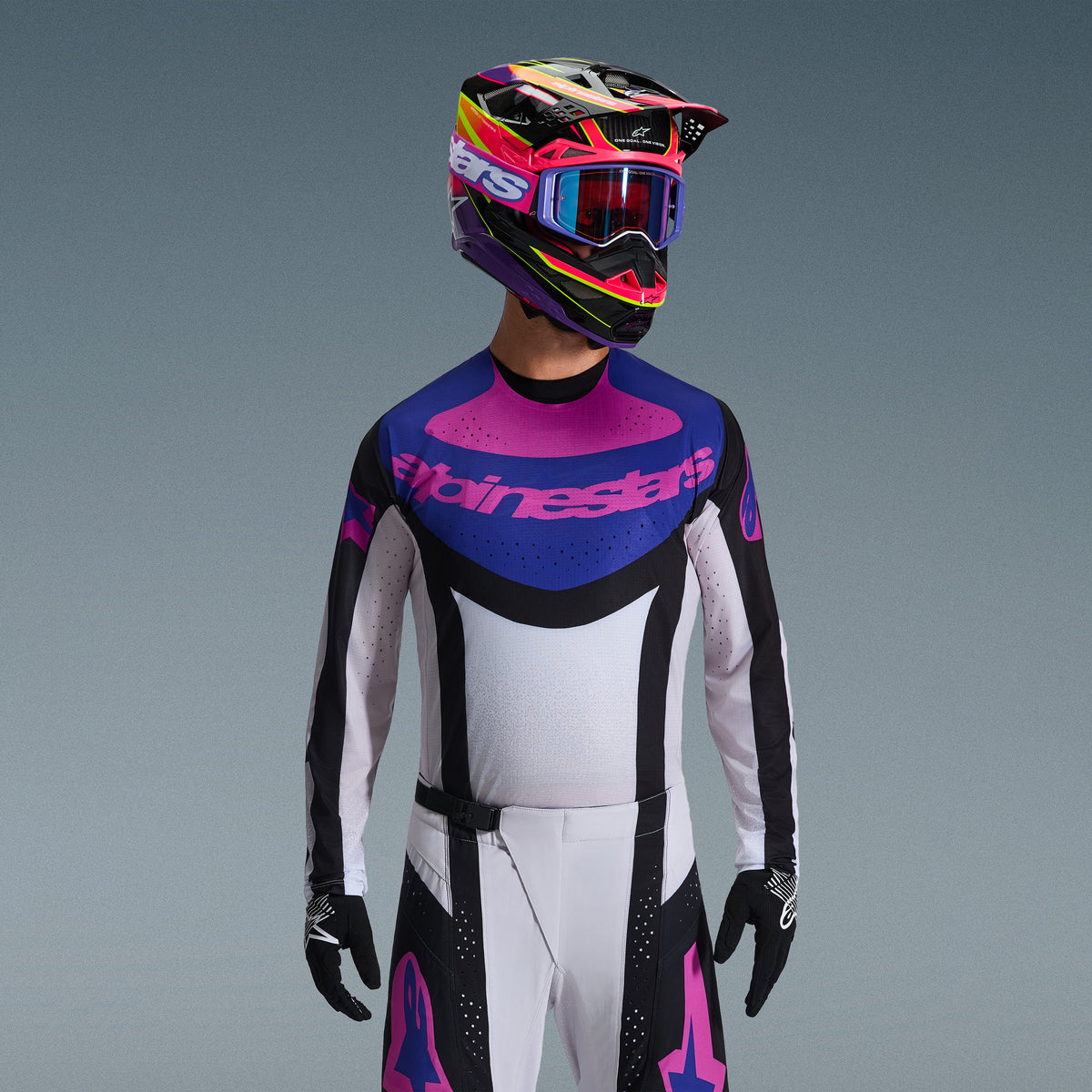 Motocrossförare i Alpinestars 2026 Techstar Knif utrustningsset, tröja med blå och rosa grafik på bröstet och vit bål, vita byxor med svarta paneler och rosa logotyper, flerfärgad hjälm i lila och rosa med matchande goggles, svarta handskar, professionell racingutrustning för terrängkörning