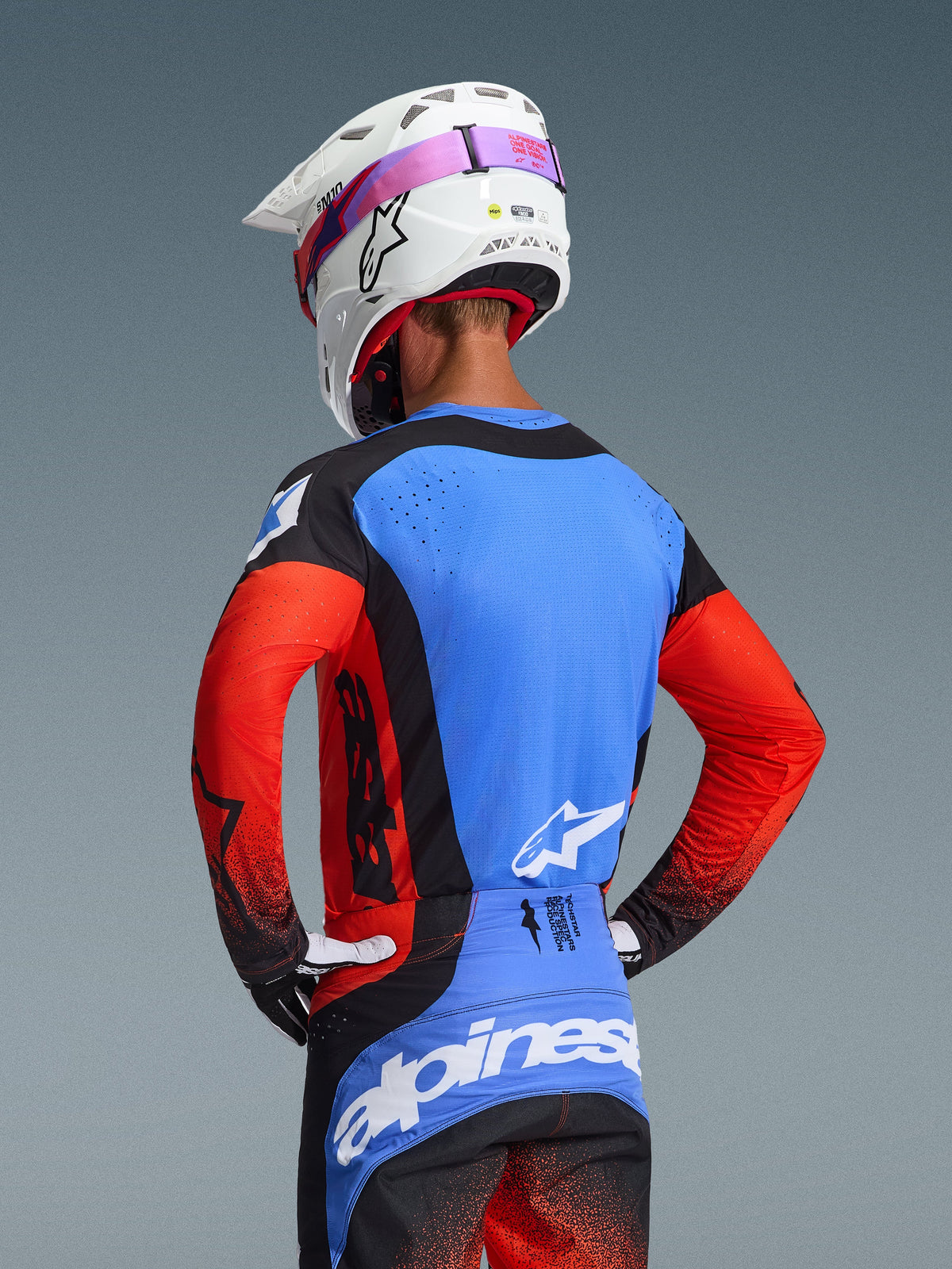 Motocross rider seen from behind wearing the 2026 Techstar Knif Gear Set, blue jersey with red sleeves and black accents, black and blue pants with white Alpinestars branding, white helmet with pink goggle strap, featuring laser-cut perforations for ventilation