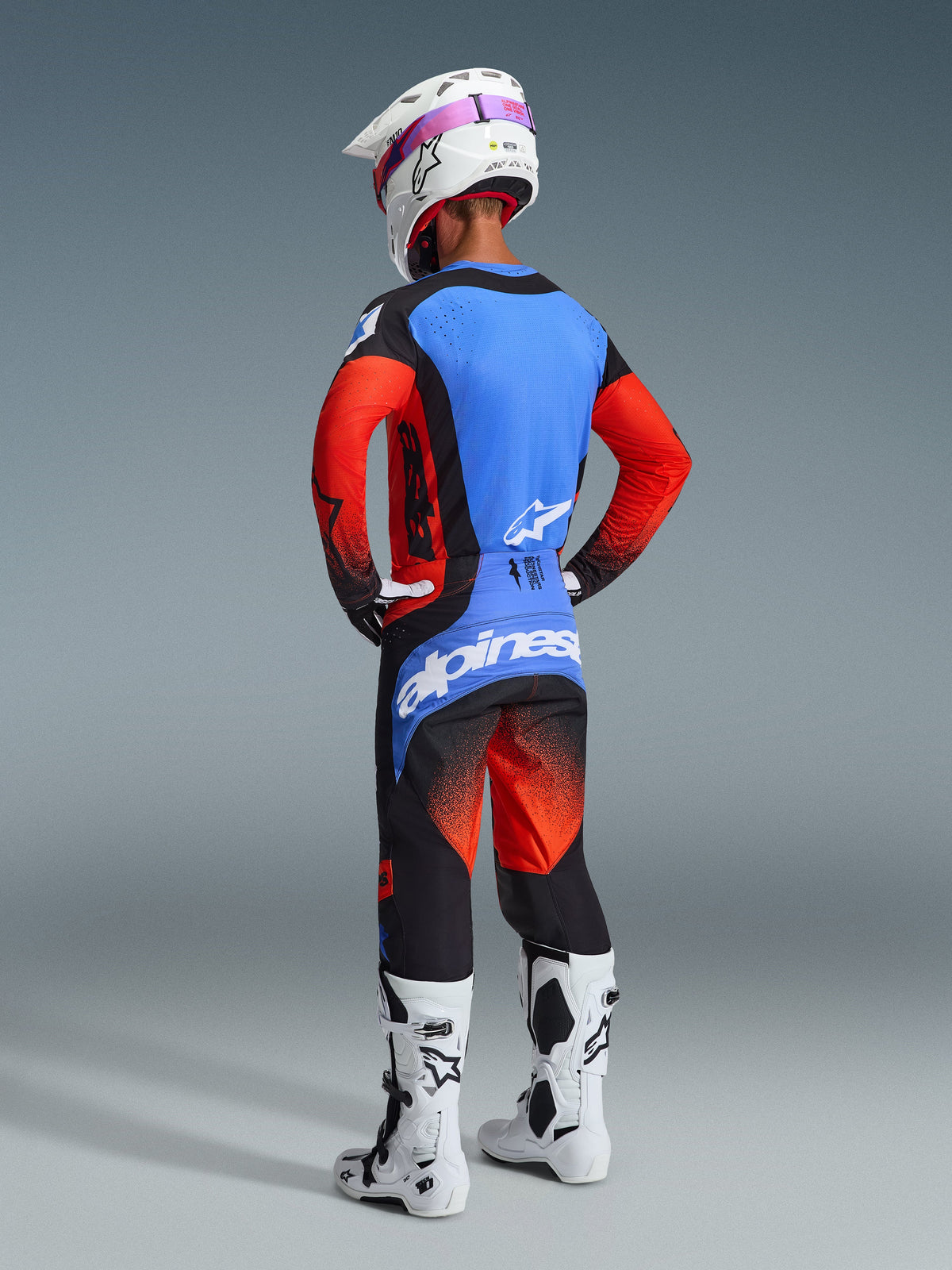 Rear view of a motocross rider wearing the Alpinestars 2026 Techstar Knif Gear Set, featuring a blue and red jersey with black accents, matching black and red pants with a large blue logo on the seat, white helmet, and white motocross boots, professional off-road racing kit