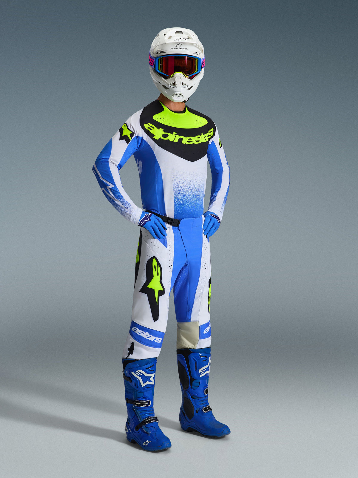 Motocross rider wearing the Alpinestars 2026 Techstar Knif Gear Set in Ucla Blue, Black, and Yellow Fluo, featuring a performance jersey, matching pants with bold branding, blue boots, blue gloves, and a white helmet with goggles, standing against a gray background.