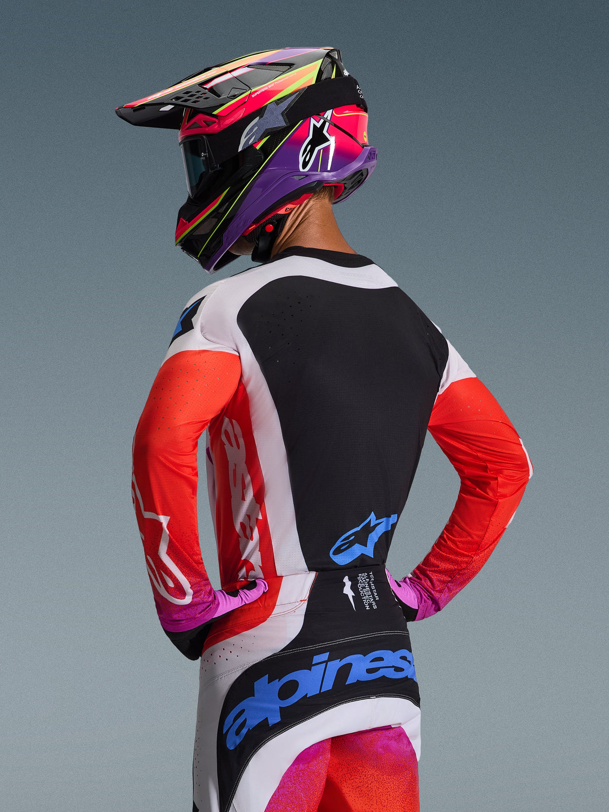 Motocrossförare sedd bakifrån klädd i Alpinestars 2026 Techstar Knif utrustningsset, neonorangea ärmar med svart ryggpanel, vita och svarta byxor med blå logotyper, livfull lila och rosa hjälm, professionell motocrossutrustning för terrängprestanda