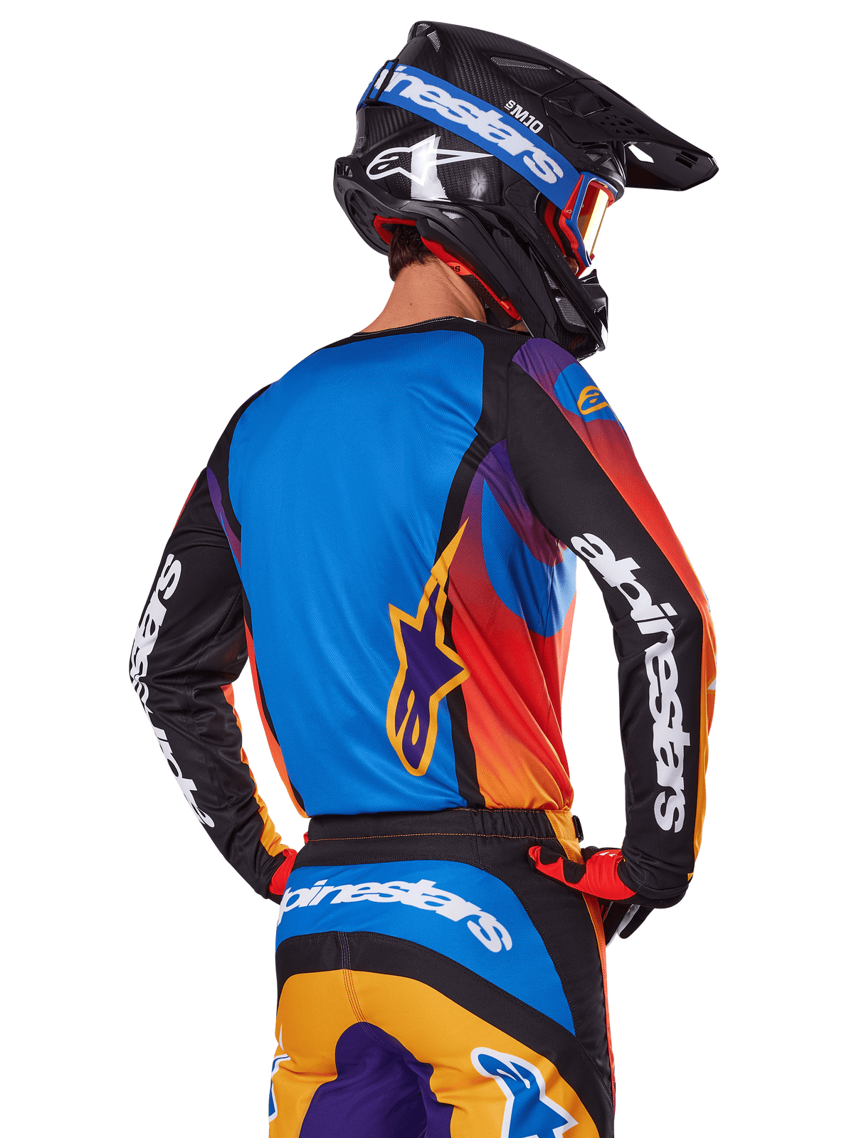 Motocrossförare sedd bakifrån som bär Alpinestars 2025 Fluid Wurx-utrustningsset, flerfärgad tröja med blå rygg och svarta ärmar med vita logotyper, matchande orangea och blå byxor, svart och blå hjälm med orangea goggles, koordinerad racingoutfit för terrängkörning.