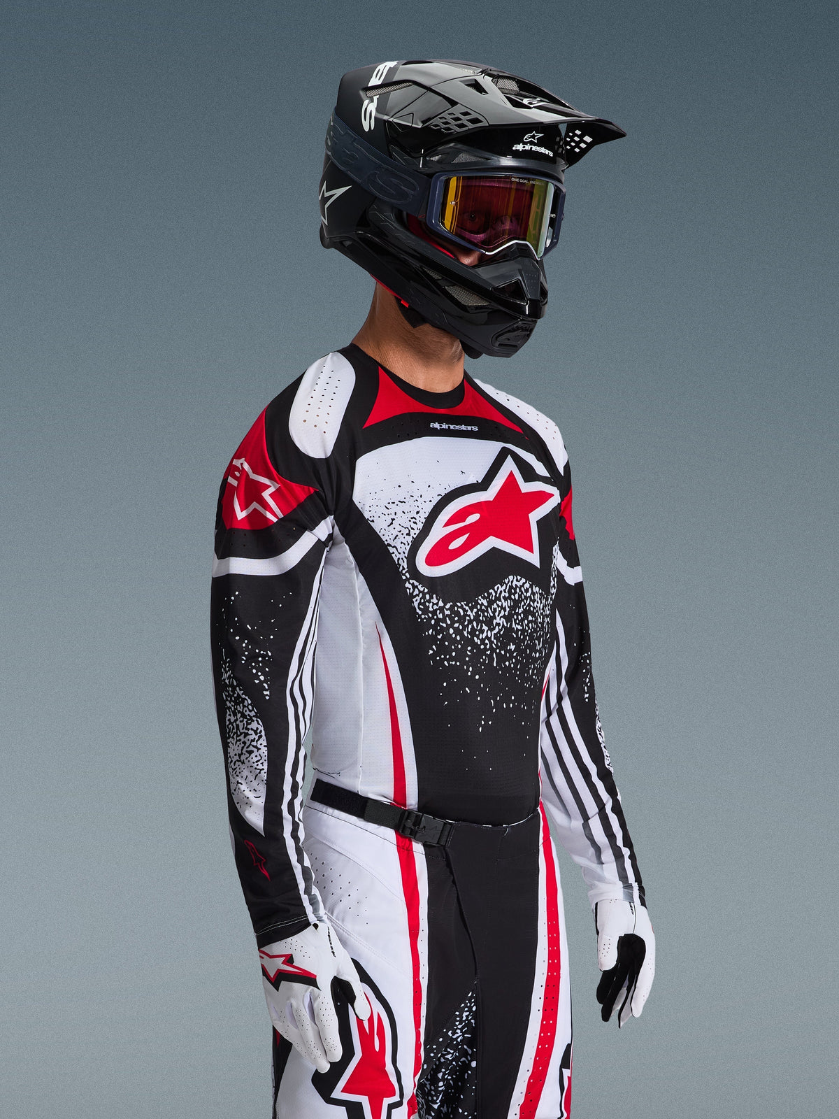 Motocrossförare som bär Alpinestars 2026 Techstar Nomur-tröja i Black White Red, med en stor röd Astars-logotyp och spräckliga grafiska mönster, kombinerat med matchande racingbyxor, vita handskar och en svart hjälm med goggles, koordinerad outfit för terrängtävling