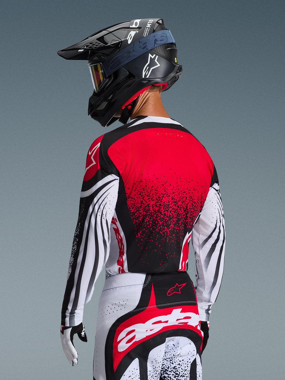 Motocrossförare sedd bakifrån bärande 2026 Techstar Nomur Jersey, röd rygg med svart gradientmönster och vita ärmar med svarta detaljer, matchande röda och vita byxor, svart hjälm och vita handskar, professionell off-road-racingutrustning