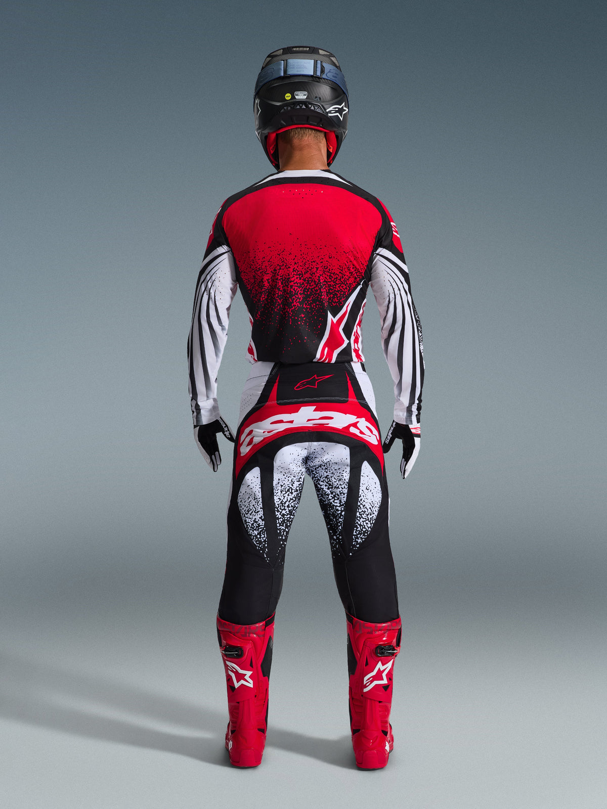 Motocrossförare sedd bakifrån i Alpinestars 2026 Techstar Nomur-utrustningsset, röd och vit tröja med svart prickigt gradientmönster, matchande vita och röda byxor med svarta detaljer, klarröda stövlar och svart hjälm, koordinerad racingutrustning för terrängkörning.