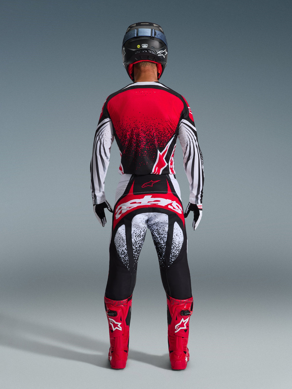 Motocrossförare sedd bakifrån i Alpinestars 2026 Techstar Nomur-tröja, röd, svart och vit med stipplat mönster, matchande racingbyxor, röda motocross-stövlar och svart hjälm, komplett utrustningsset för terrängkörning