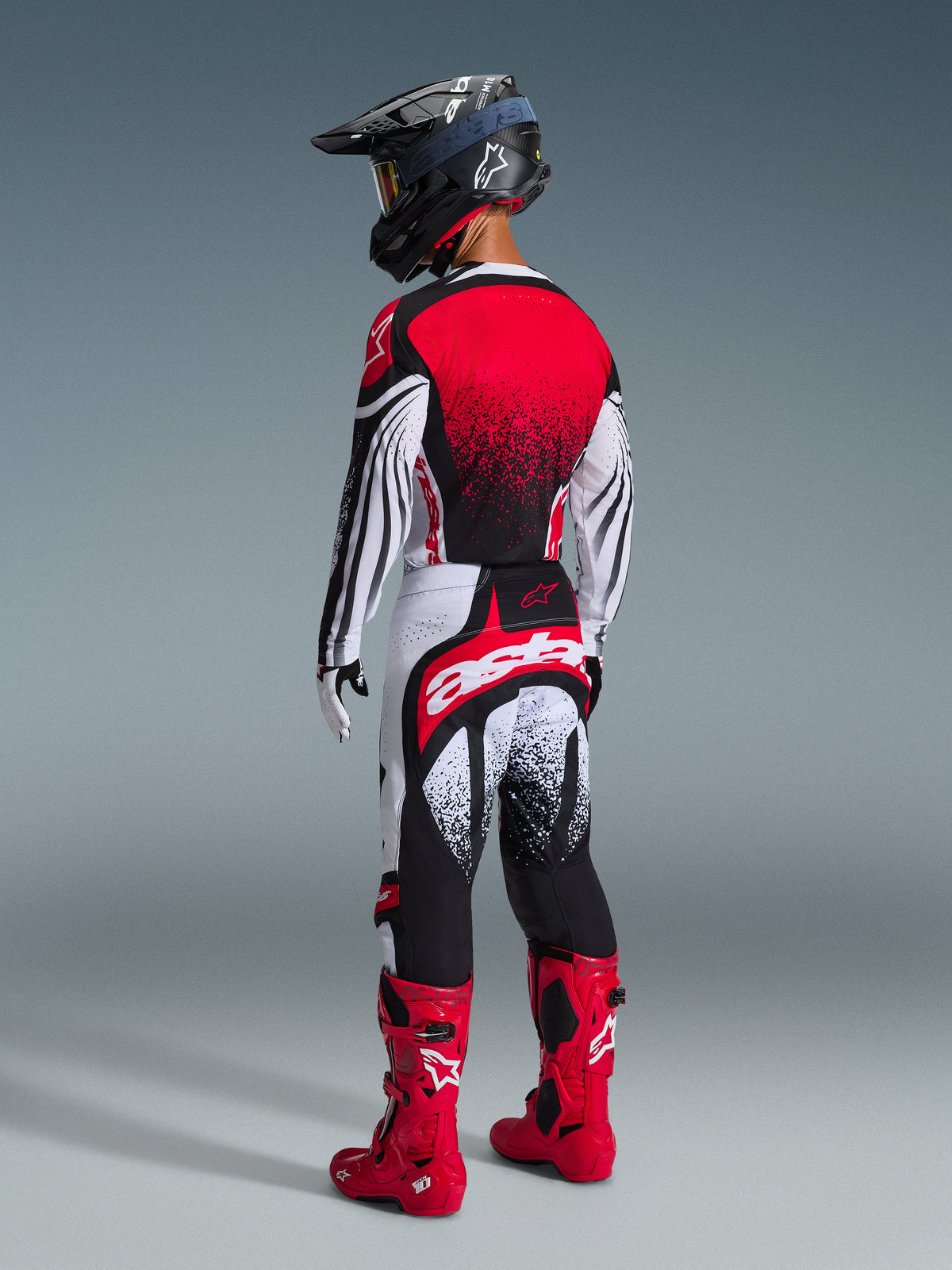 Motocrossförare sedd bakifrån i Alpinestars 2026 Techstar Nomur-utrustningsset, röd, vit och svart tröja med prickig gradient, matchande byxor med framträdande 'astars'-logotyp, svart hjälm och röda motocross-stövlar, professionell utrustning för terrängkörning
