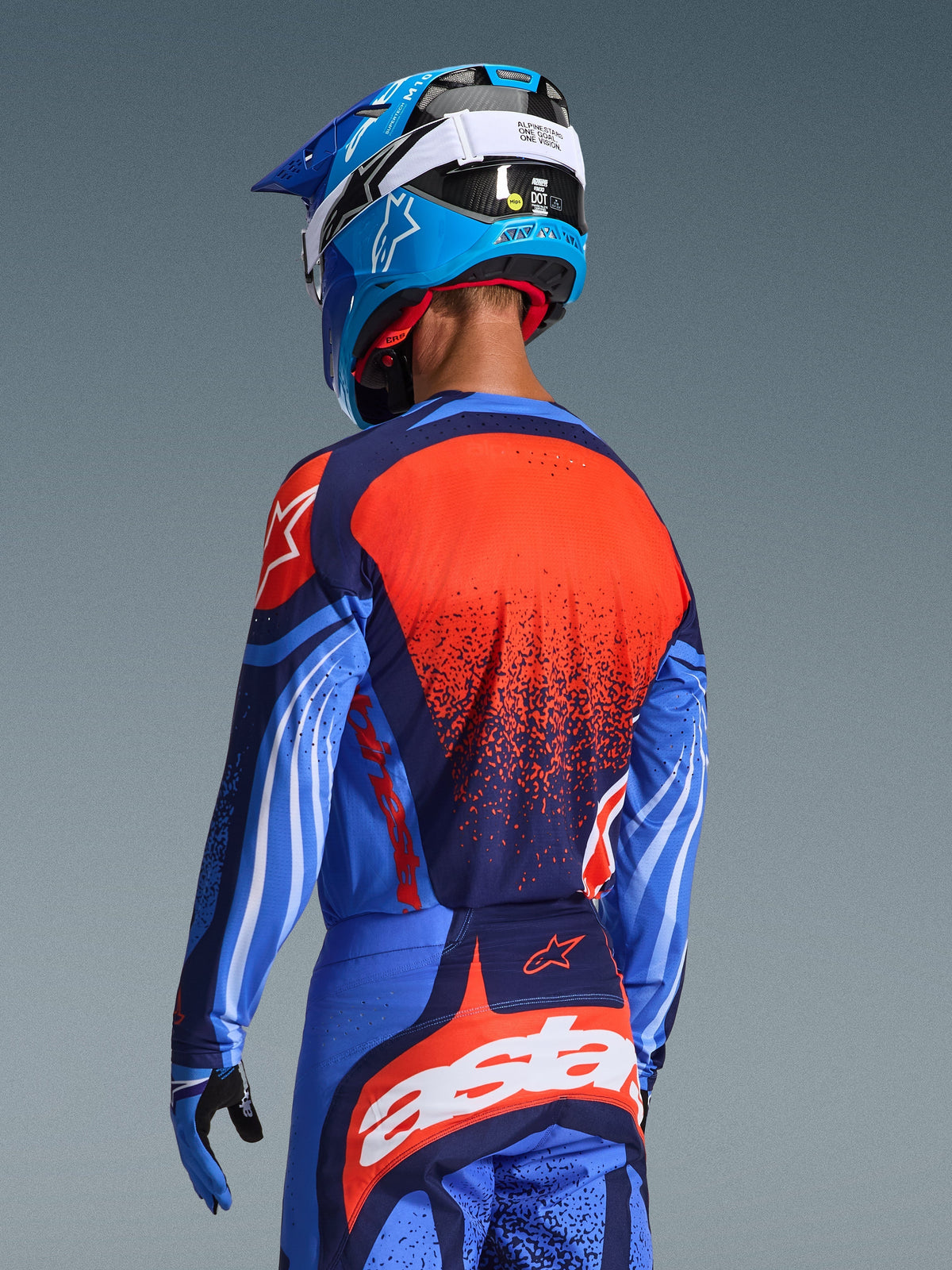 Motocrossförare sedd bakifrån iklädd 2026 Techstar Nomur-tröja, mörkblå med orangea och UCLA-blå detaljer, med en stor orange texturerad grafik på ryggen, matchad med koordinerande motocrossbyxor och en blå racinghjälm, professionell utrustning för terrängkörning.