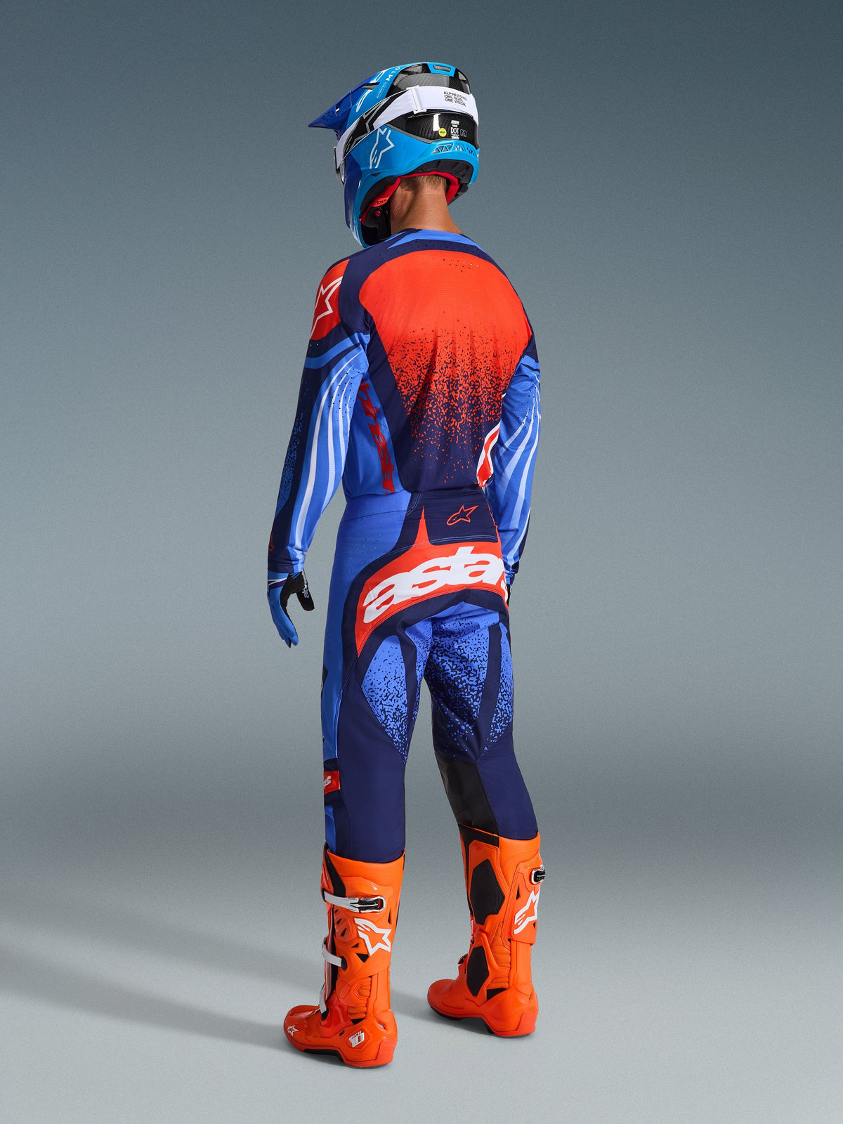 Motocrossförare sedd bakifrån i Alpinestars 2026 Techstar Nomur-tröja i Dark Navy, Orange och UCLA Blue, matchande byxor med tydlig vit logotyp, orangea stövlar och blå-vit hjälm, komplett utrustning för terrängkörning.
