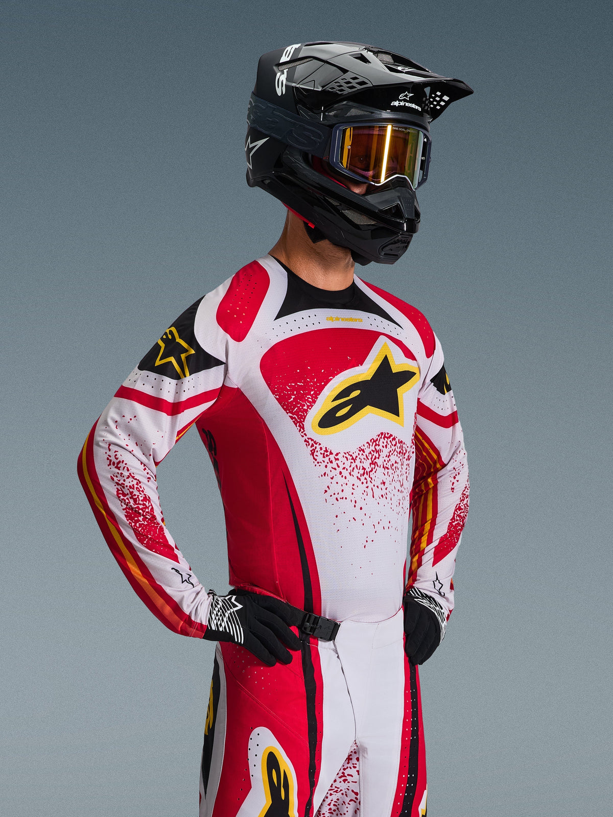 Motocrossförare i Alpinestars 2026 Techstar Nomur-tröja, röd, vit och svart med gula detaljer, matchande byxor, svart hjälm med guldfärgade goggles och svarta handskar, atletisk tävlingspassform för professionell terrängkörning
