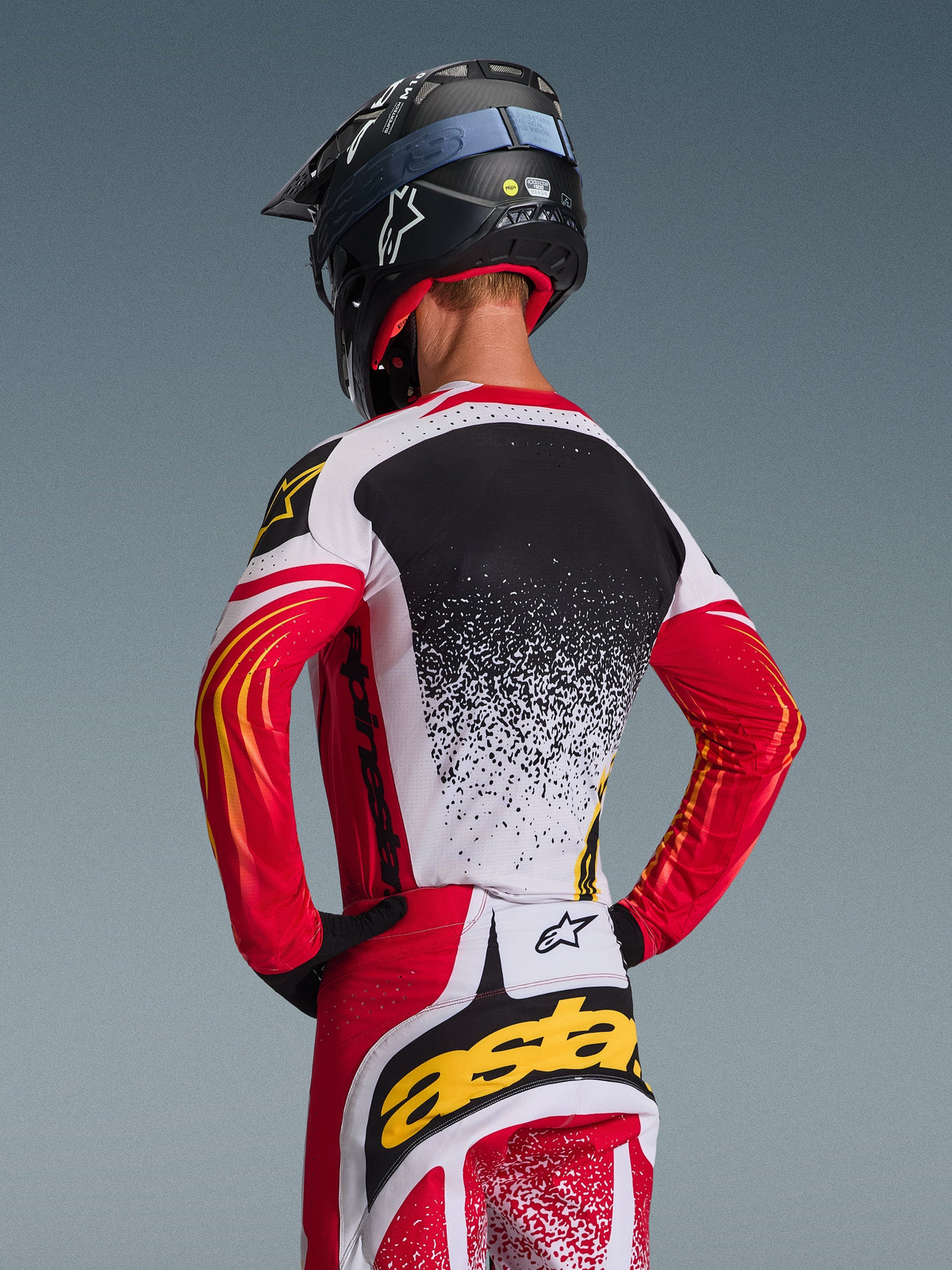 Motocrossförare bakifrån i Alpinestars 2026 Techstar Nomur-tröja och matchande byxor, röd, vit och svart med gula detaljer, med en spräcklig gradientdesign på ryggen, svart Alpinestars-hjälm och svarta handskar, professionell utrustning för terrängkörning