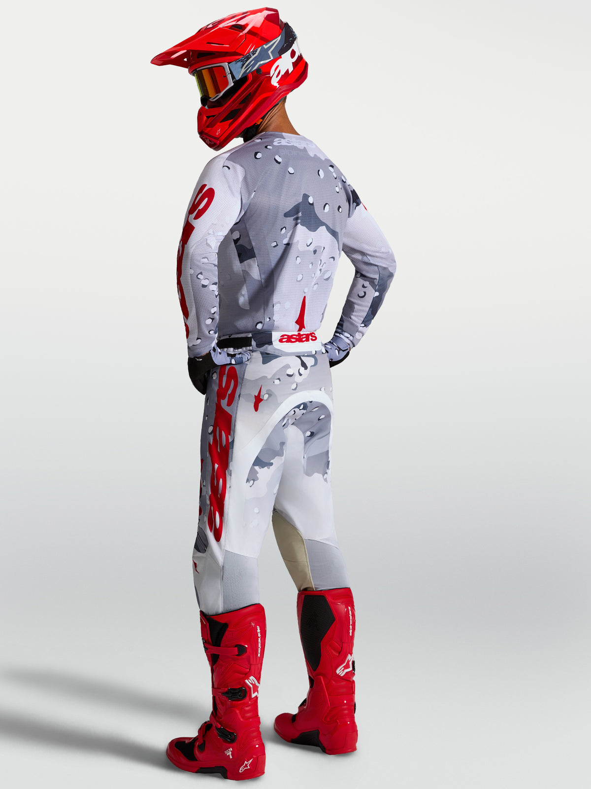 Alpinestars Racer Air Lone tröja och motocross-ställ i grå camo och rött, andningsbar meshkonstruktion för terrängkörning, visas bakifrån på modell med röda stövlar och hjälm.