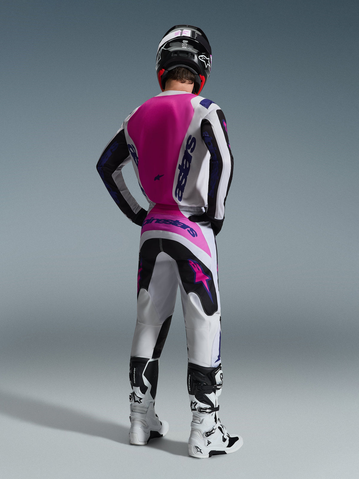 Motocross rider from the back wearing Alpinestars 2026 Fluid Grid Jersey, white jersey with large hot pink back panel and purple branding, matching white and black pants with pink accents, white boots, and black helmet, coordinated off-road racing gear