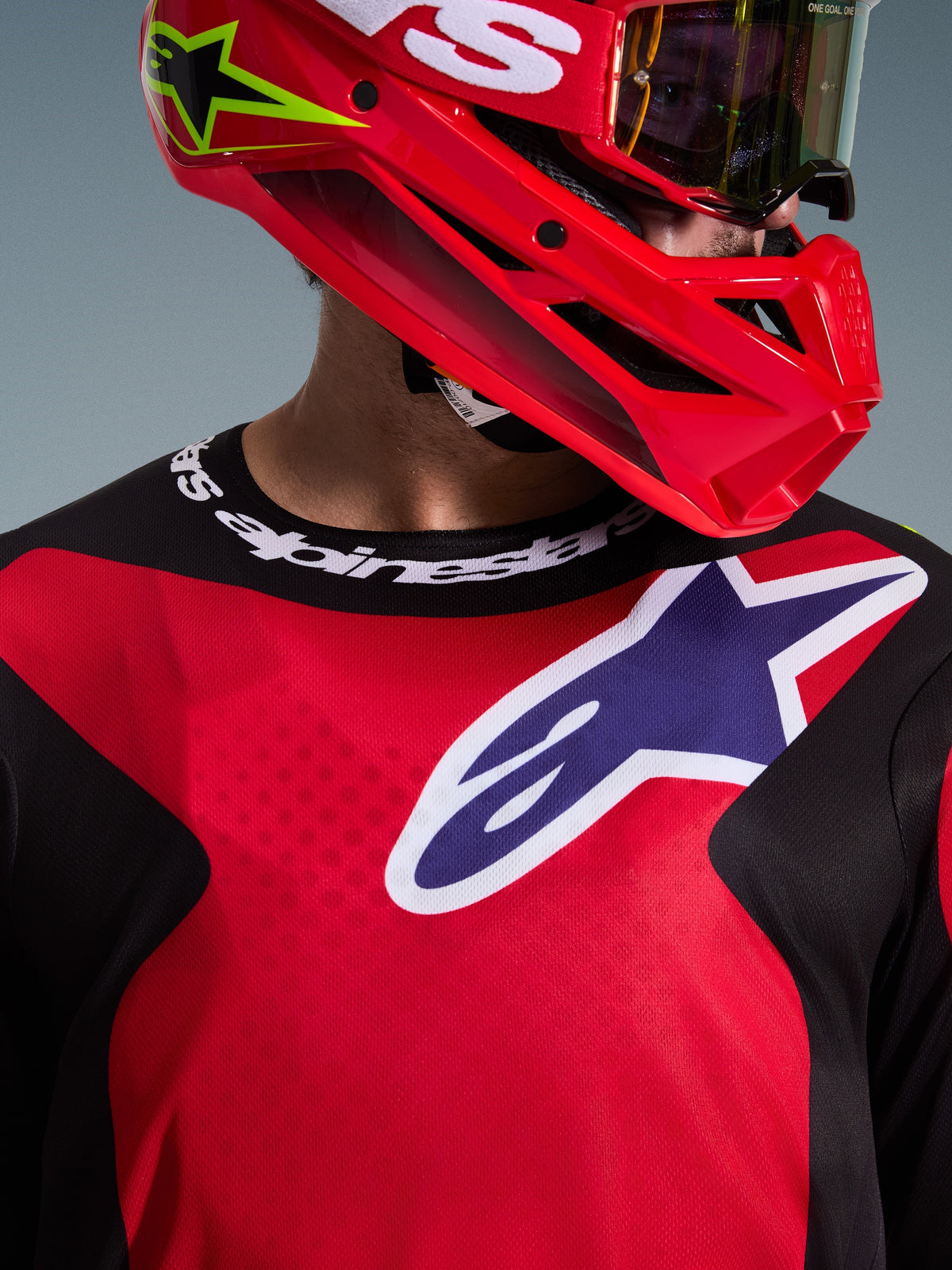Close-up of a motocross rider wearing the 2026 Fluid Grid Jersey in Red, Black, and Purple, featuring a large Alpinestars logo on the chest and branded sport collar, paired with a red helmet and goggles, highlighting the performance polyfabric and athletic fit.