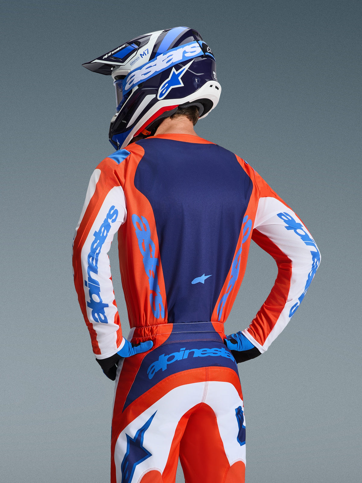 Alpinestars 2026 Fluid Grid Jersey, vy bakifrån av motocrossförare i matchande orange och UCLA-blått utrustningsset, marinblå ryggpanel, vita ärmdetaljer med blå branding, matchande hjälm, högpresterande terrängkläder